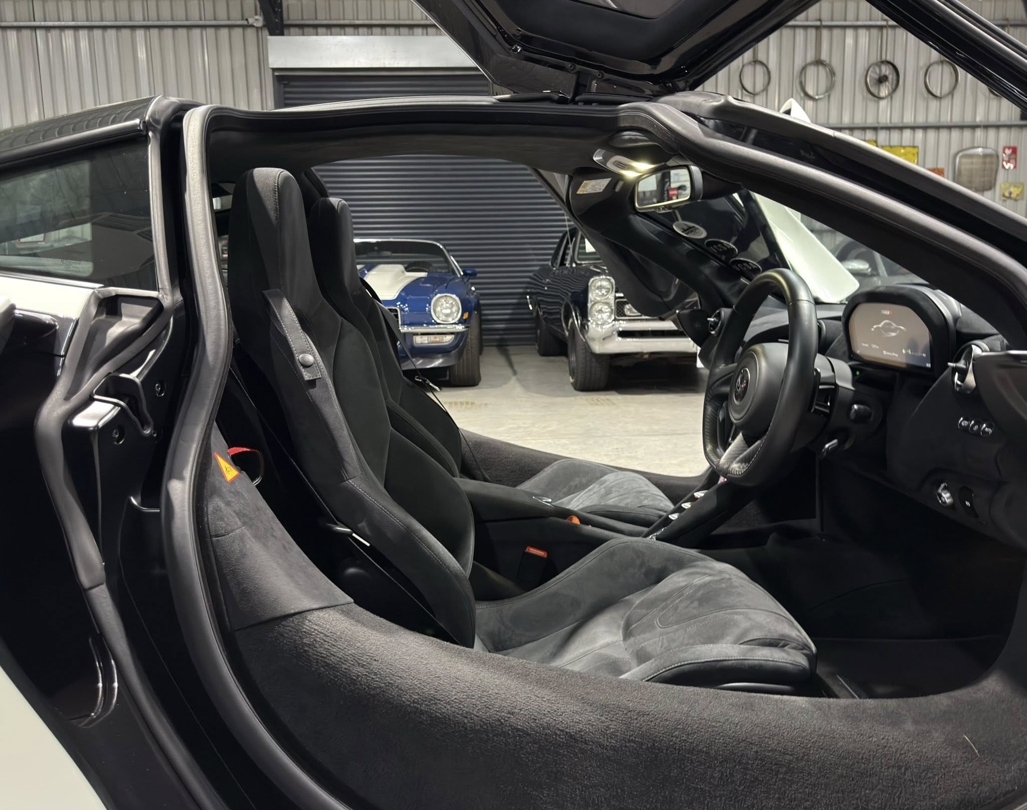 Interior of a black convertible with seats visible, roof retracted, and other classic cars in a garage behind.