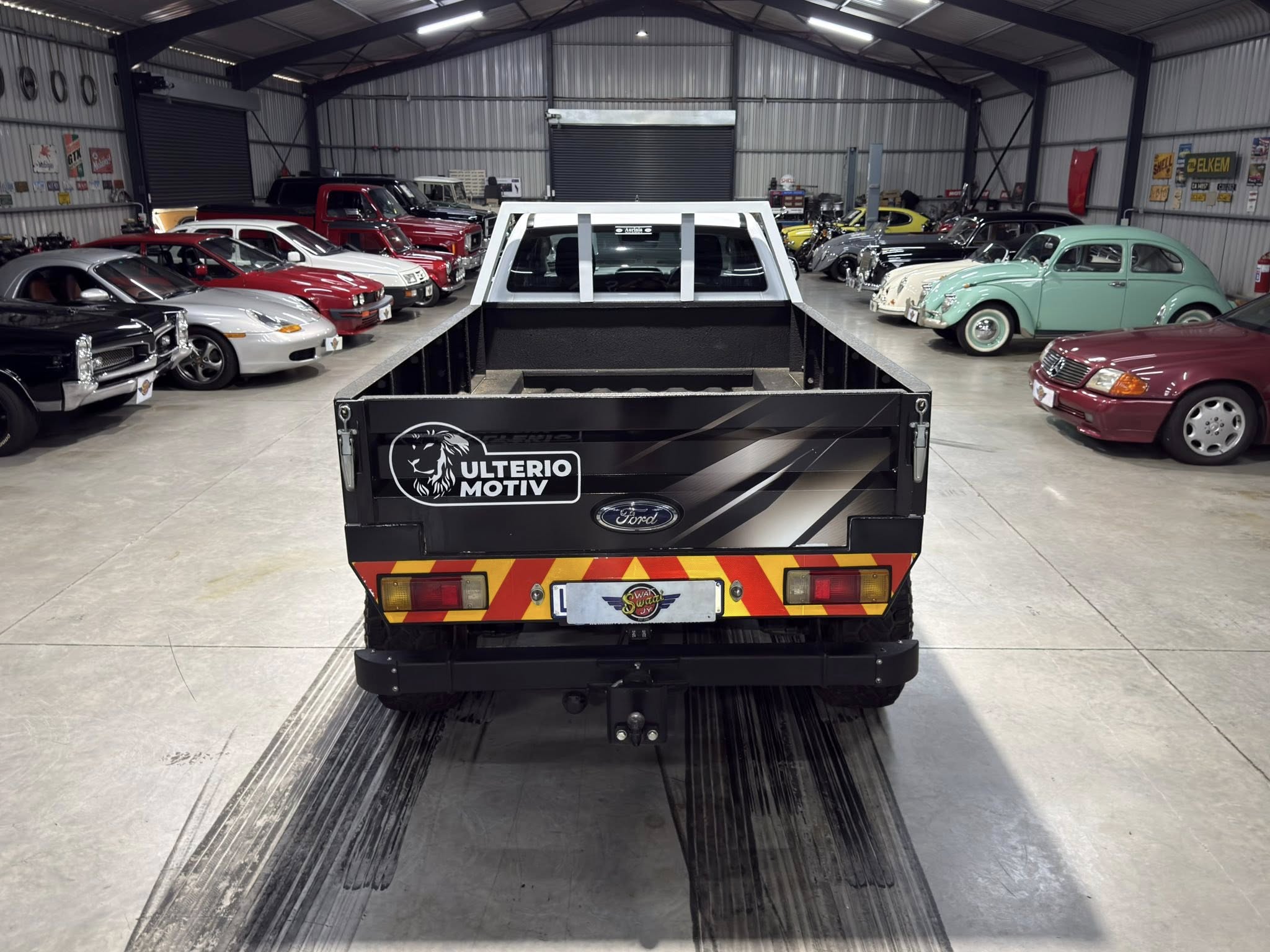 Indoor showroom-style garage filled with vintage cars parked along both sides, a black branded trailer in the foreground center, and a metal interior.