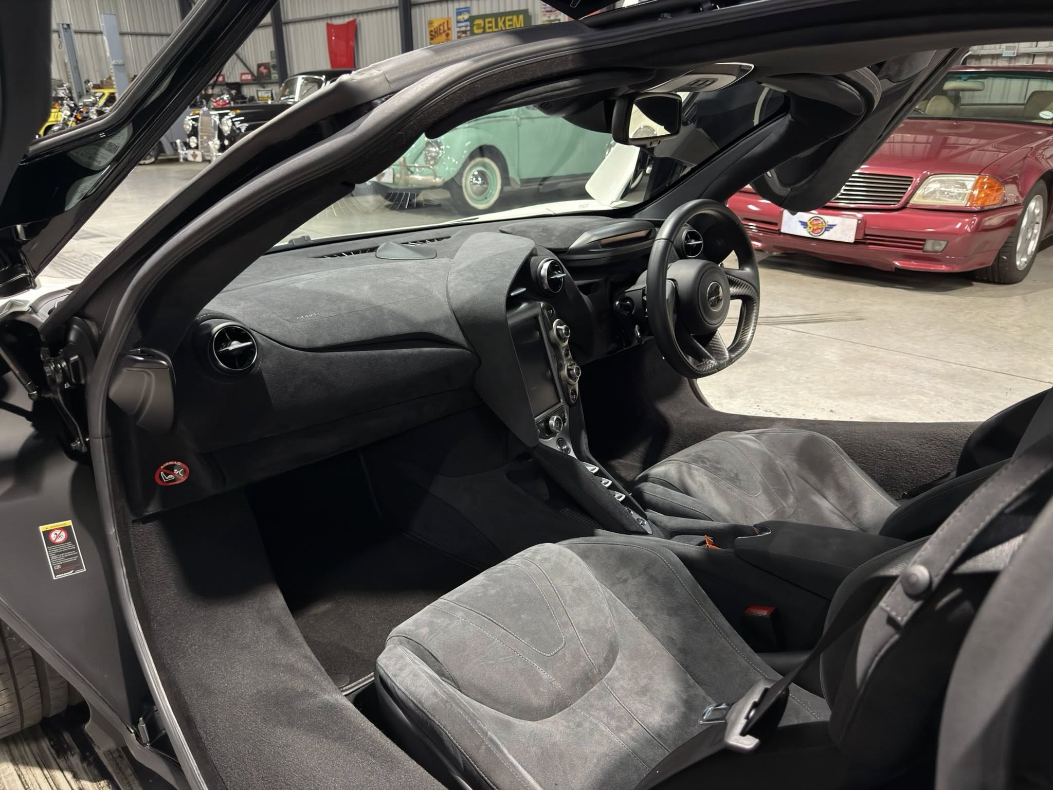 Open-door view of a black luxury coupe interior with dashboard, steering wheel, and center console; vintage cars in a showroom backdrop.