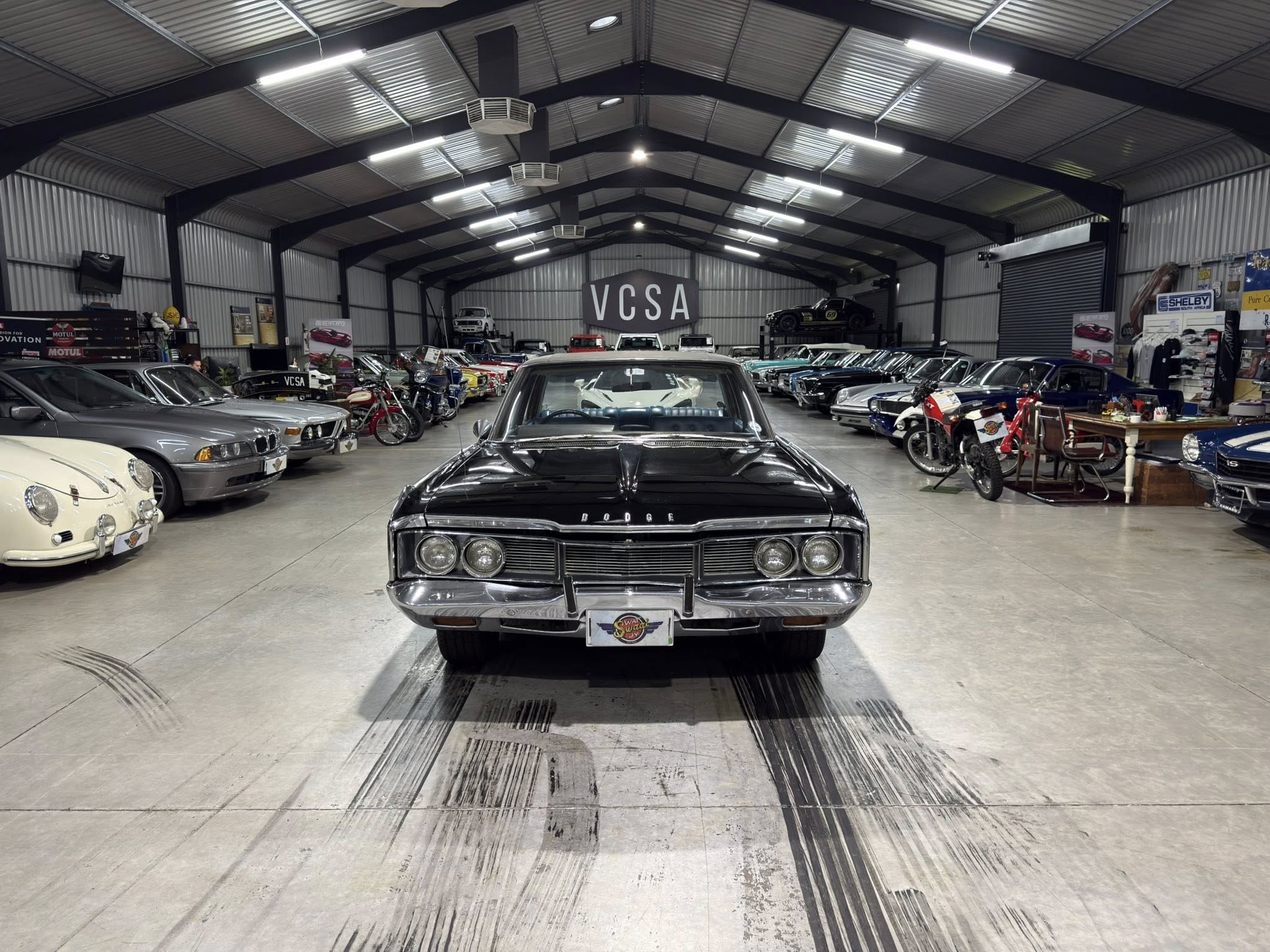 Indoor classic car showroom with a row of vintage cars on either side and a black Dodge in the foreground; VCSA sign visible at the back.