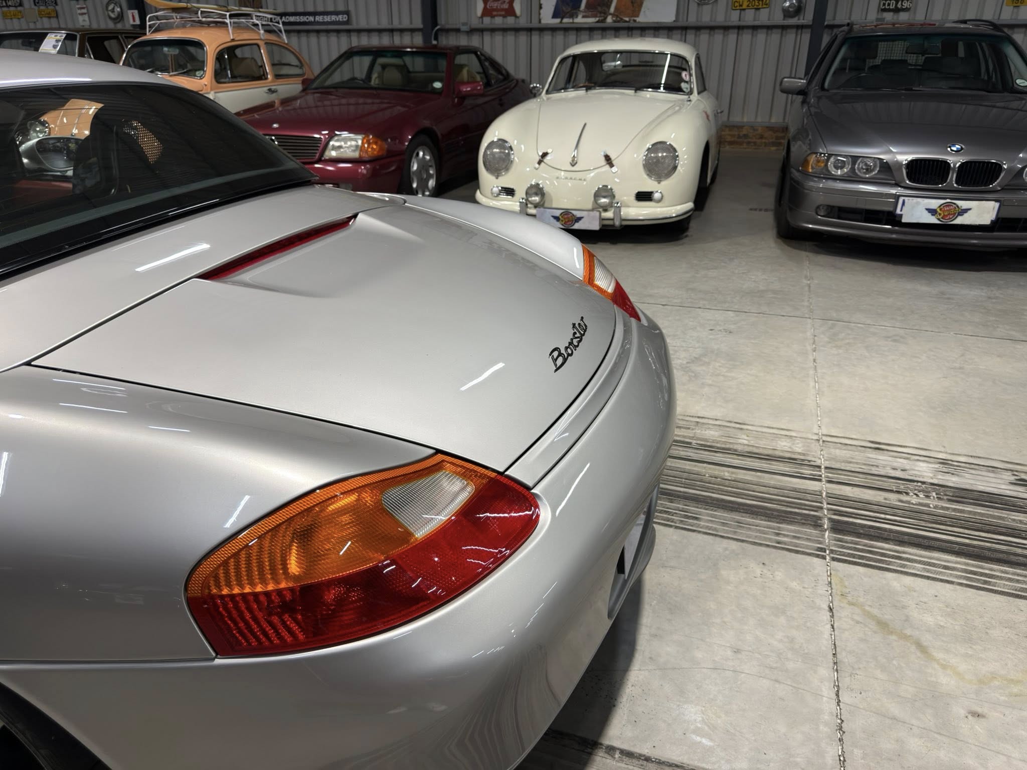 Rear view of a silver Porsche with orange taillights in a showroom filled with classic and modern cars in the background.