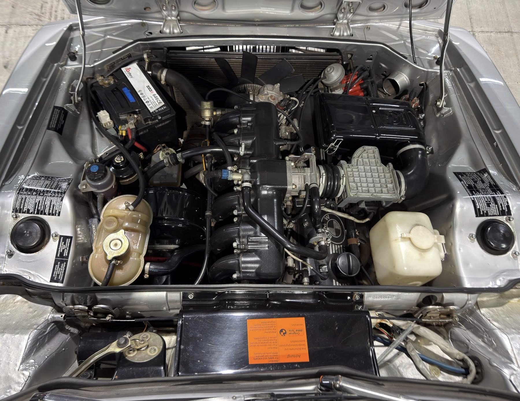Open car hood showing engine bay with battery, air intake, radiator, and coolant bottle.