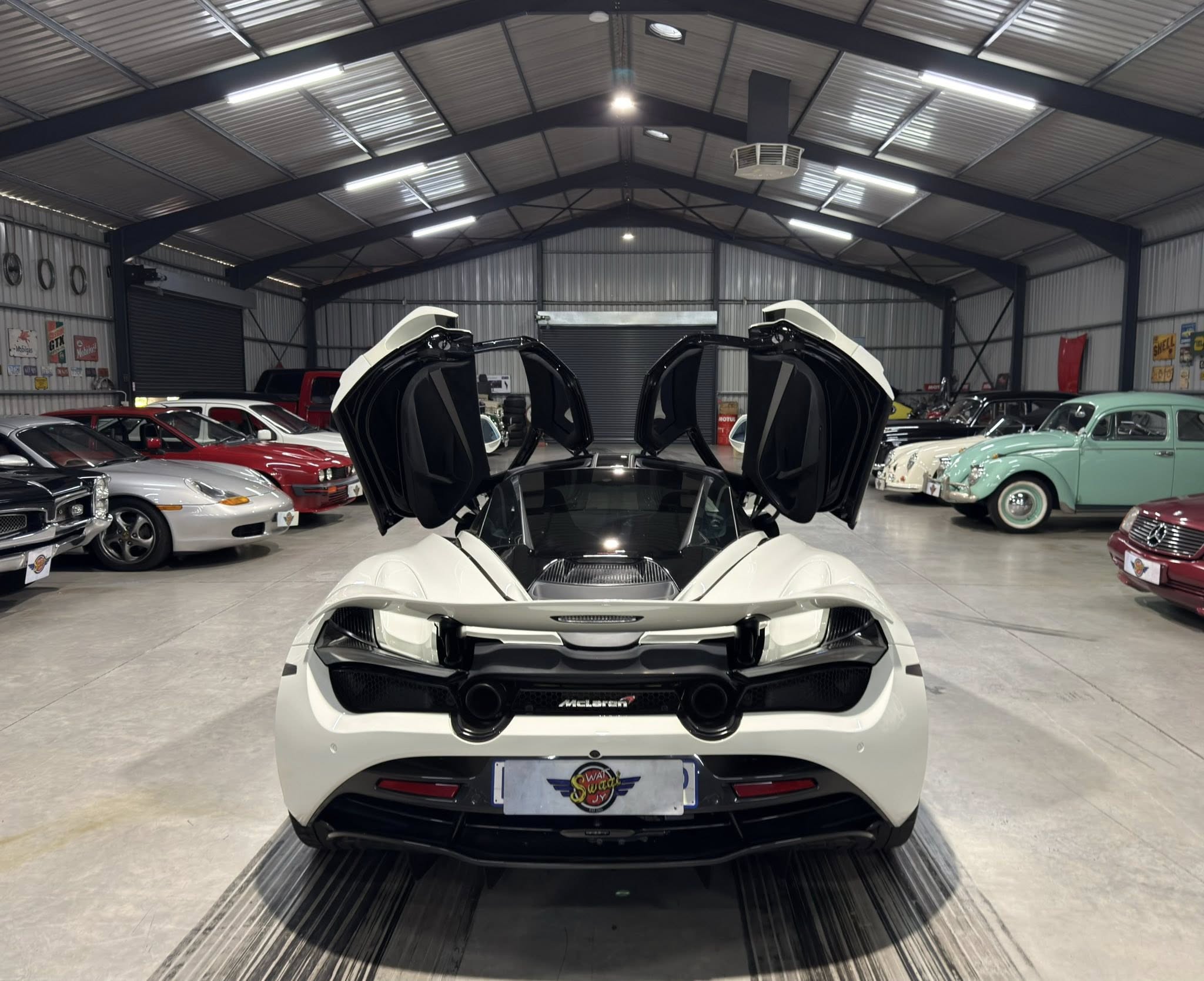 White McLaren with gull-wing doors open in a spacious showroom surrounded by classic cars
