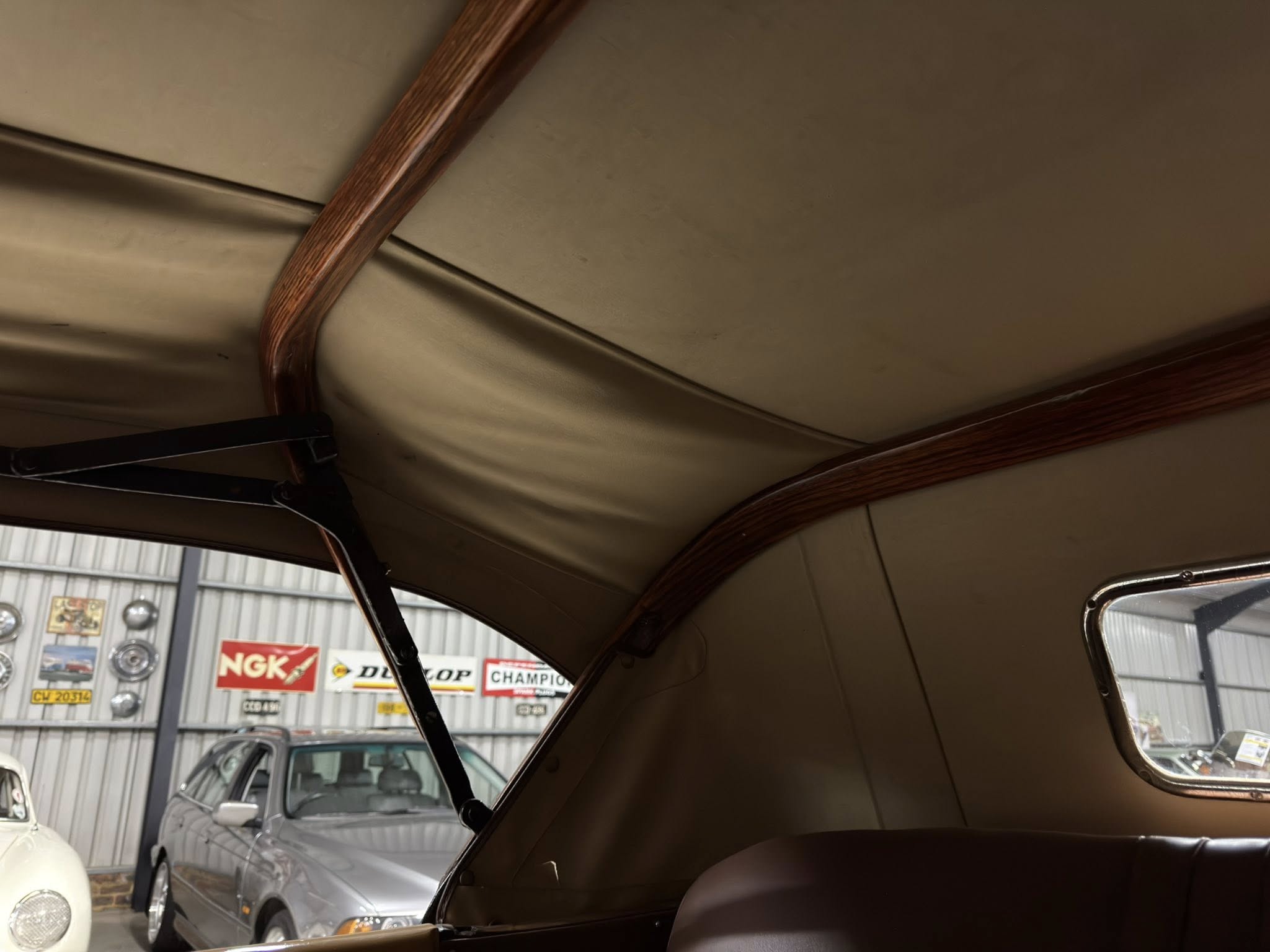Interior of a vintage car showing brown leather and wood trim, with a showroom and classic cars visible outside through windows.