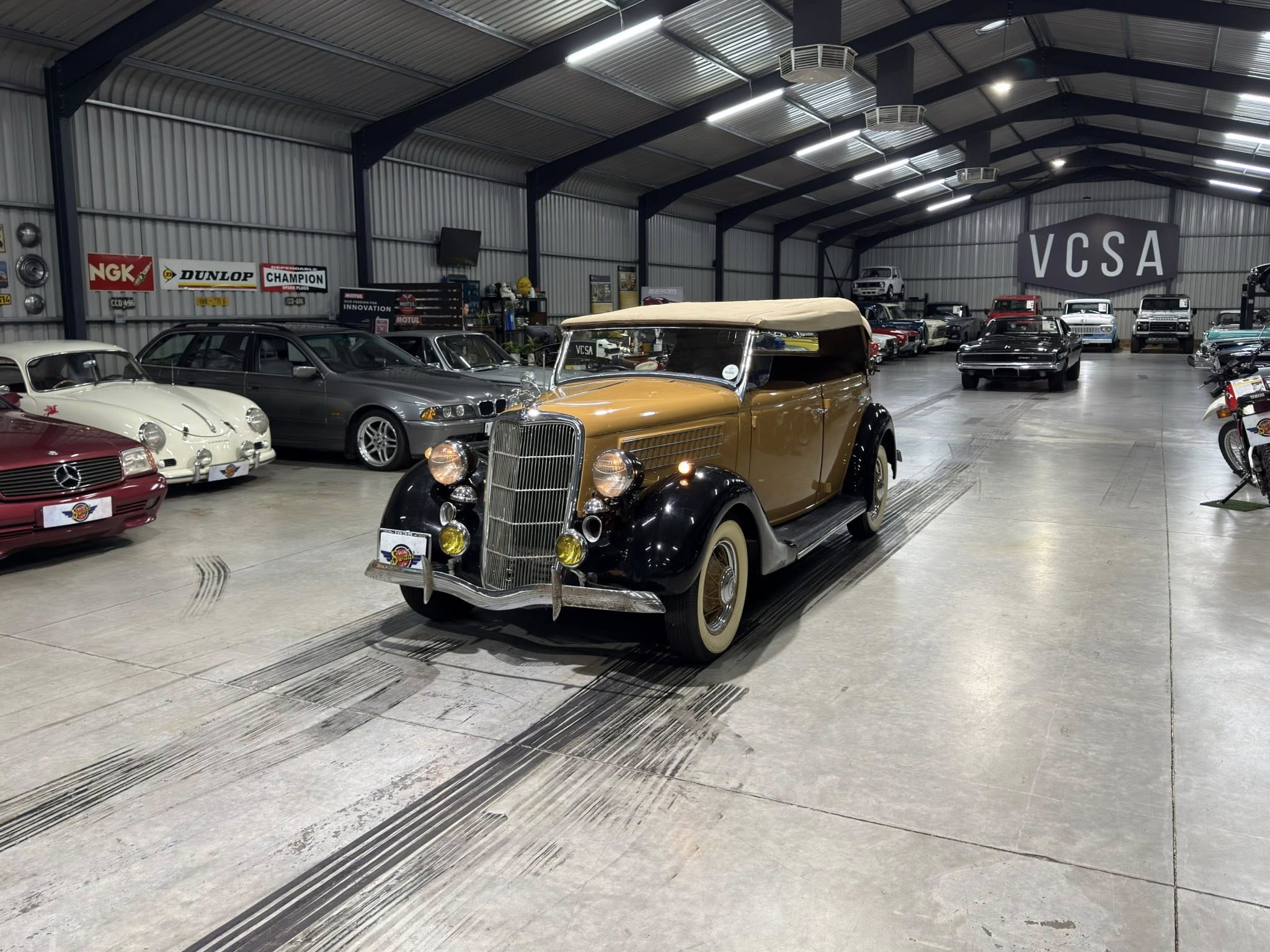 Indoor showroom filled with vintage cars; beige convertible in front and a large VCSA sign in the background