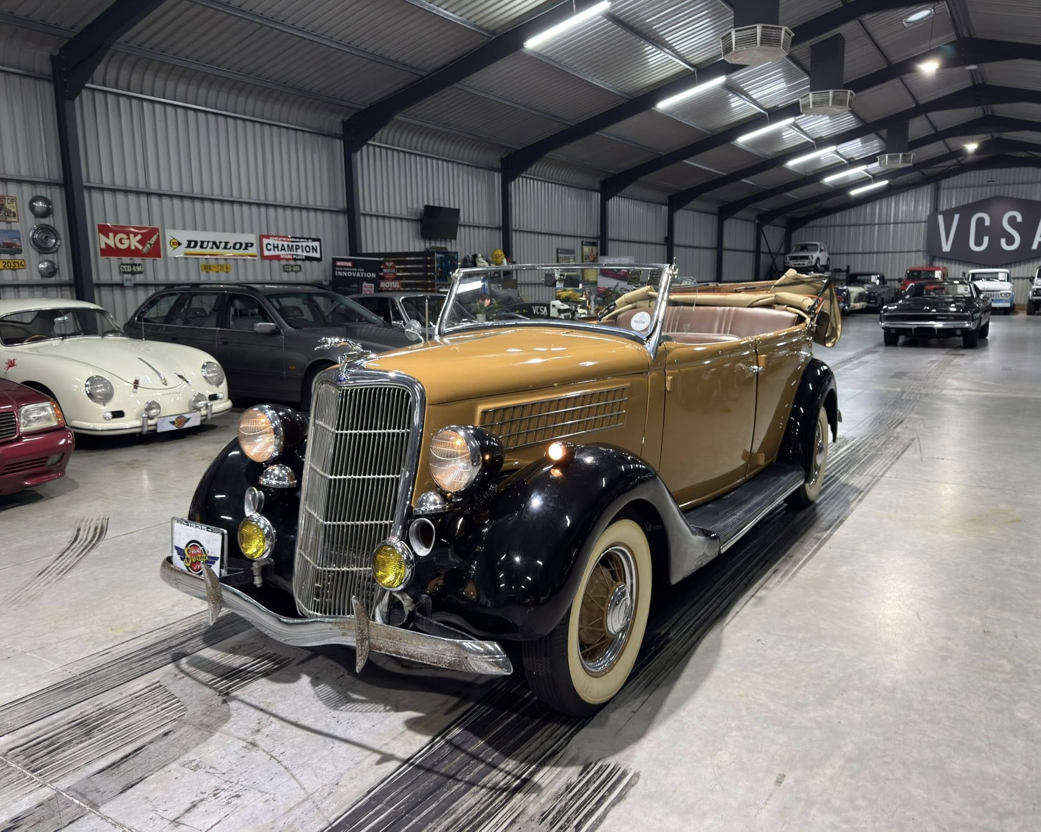 Inside a large industrial-style showroom with vintage cars, a tan convertible leads the row of classic vehicles in the foreground.