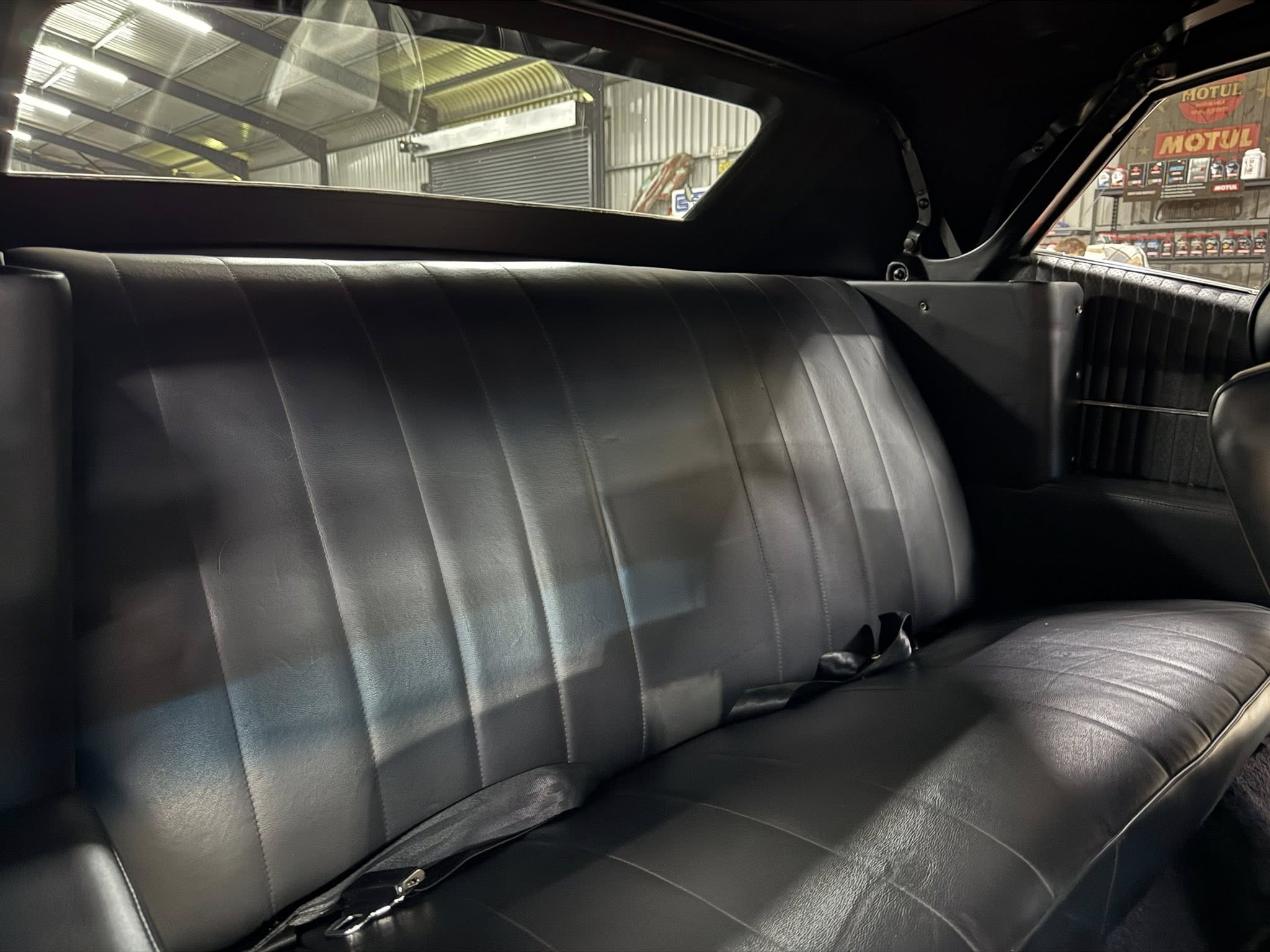 Black leather car back seat with seat belts, view through rear window of a workshop with Motul banners outside