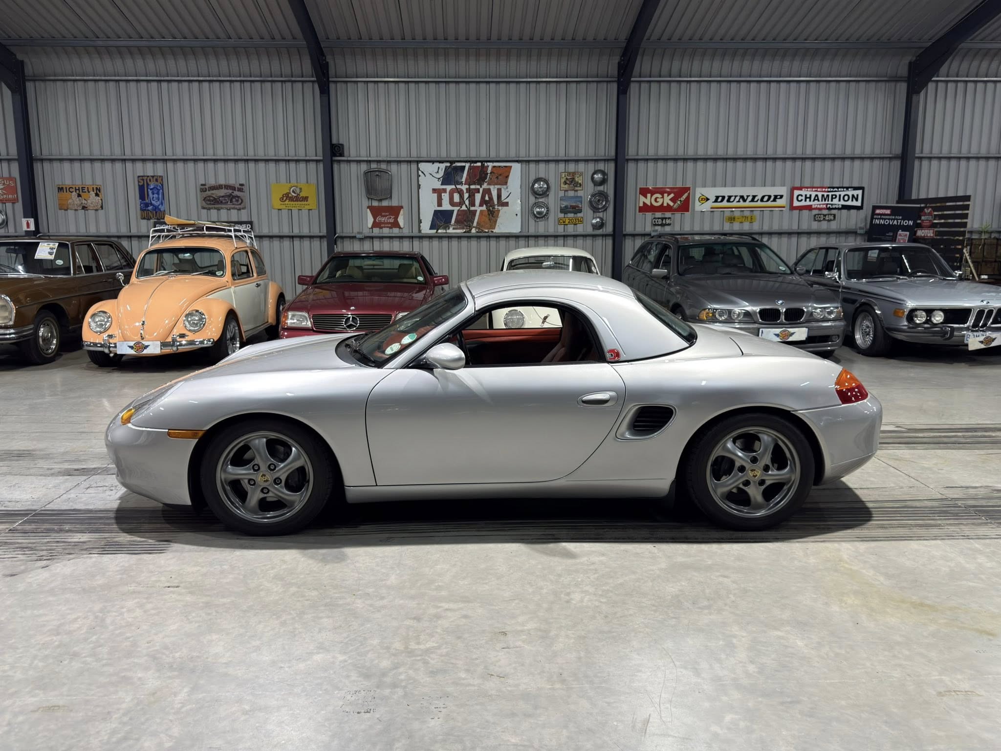 Indoor garage showroom with a row of classic cars; foreground features a silver sports car, orange vintage Beetle to the left, and posters on the corrugated metal wall behind them.