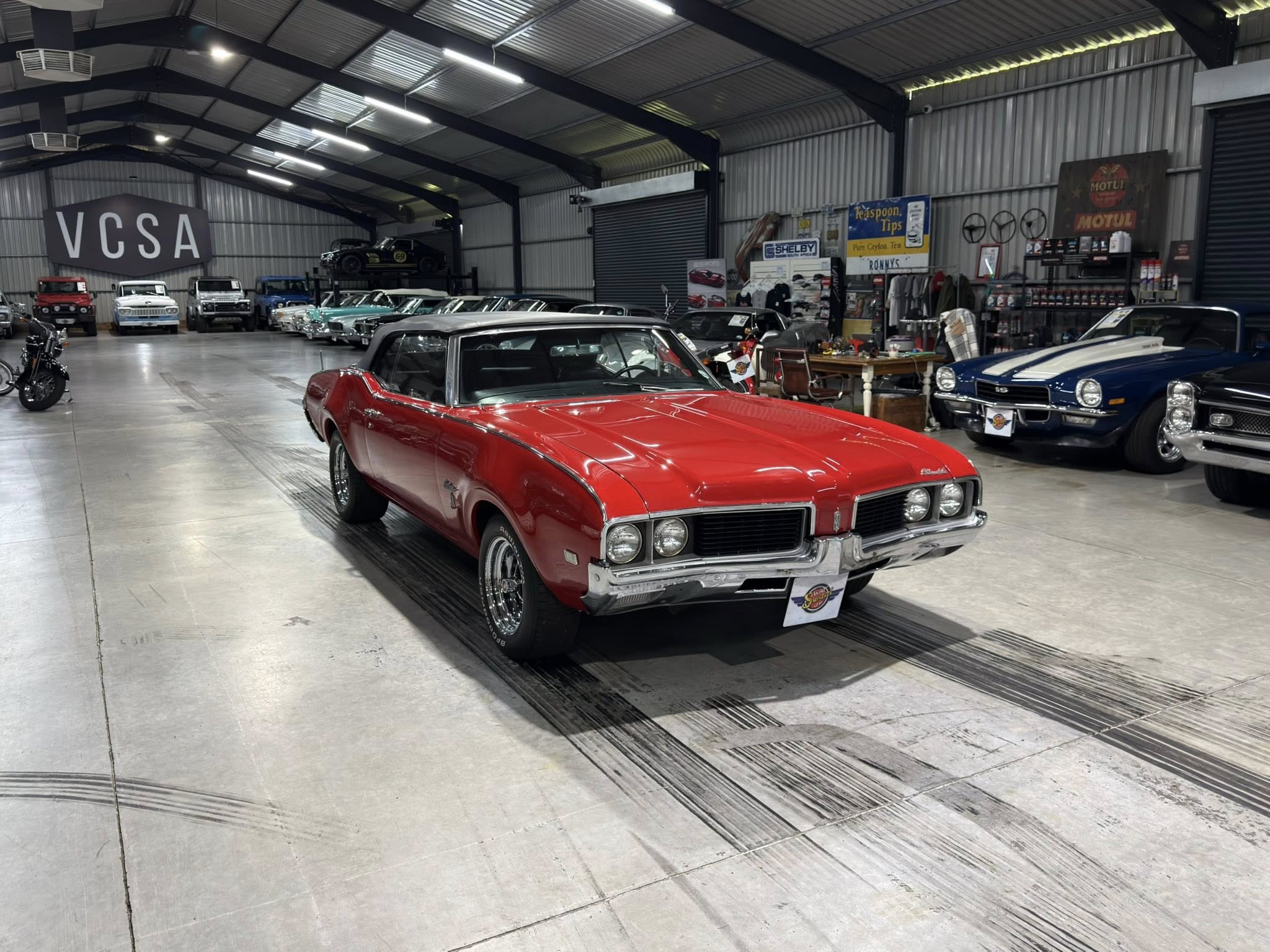 Red vintage convertible in the foreground of a large indoor classic car showroom, with more classic cars lined up along the sides and a VCSA sign on the left.