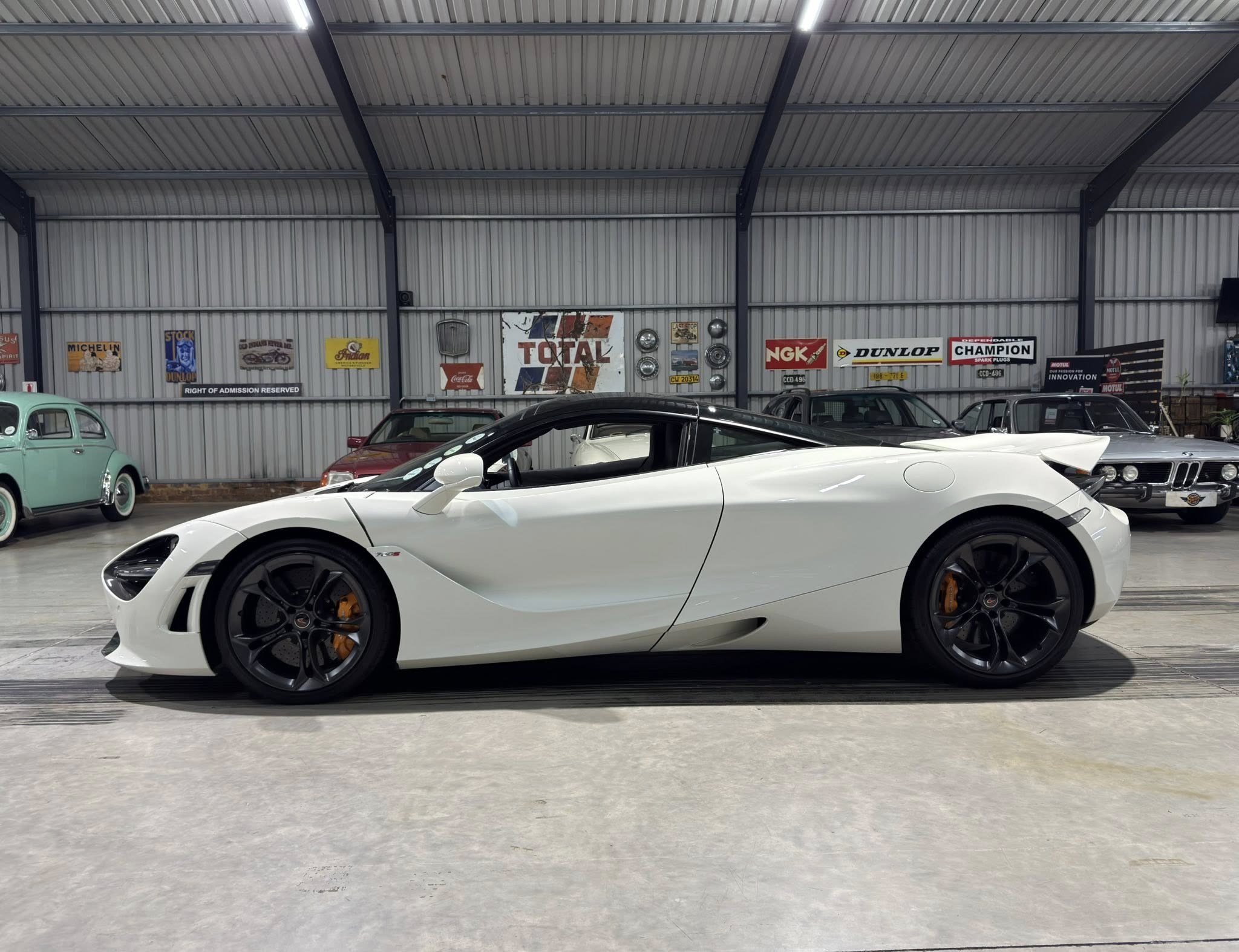 White sports car in a showroom with vintage signs on the wall and several other classic cars in the background.