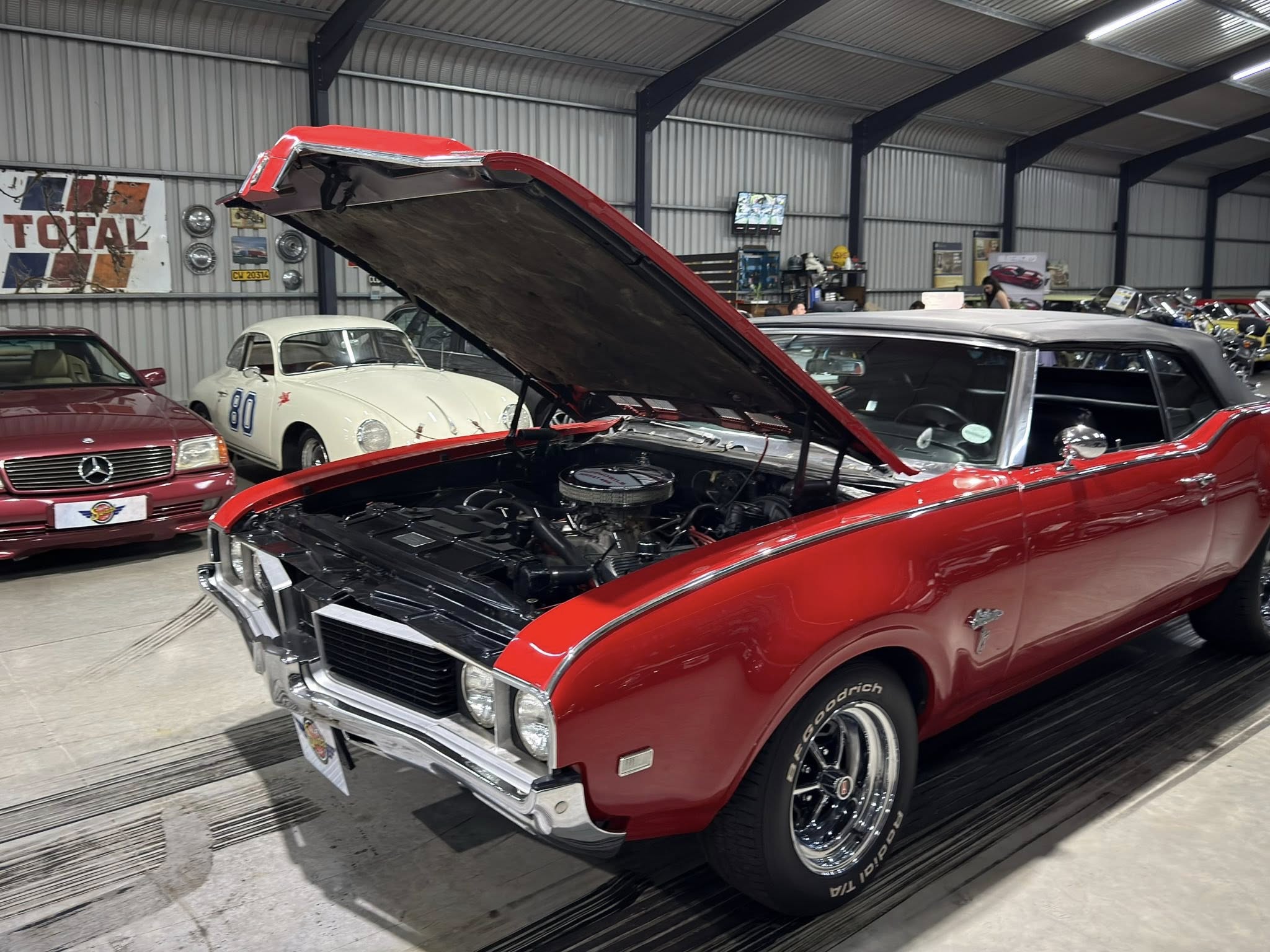 Red vintage car with hood open inside a showroom filled with classic cars and retro decor.