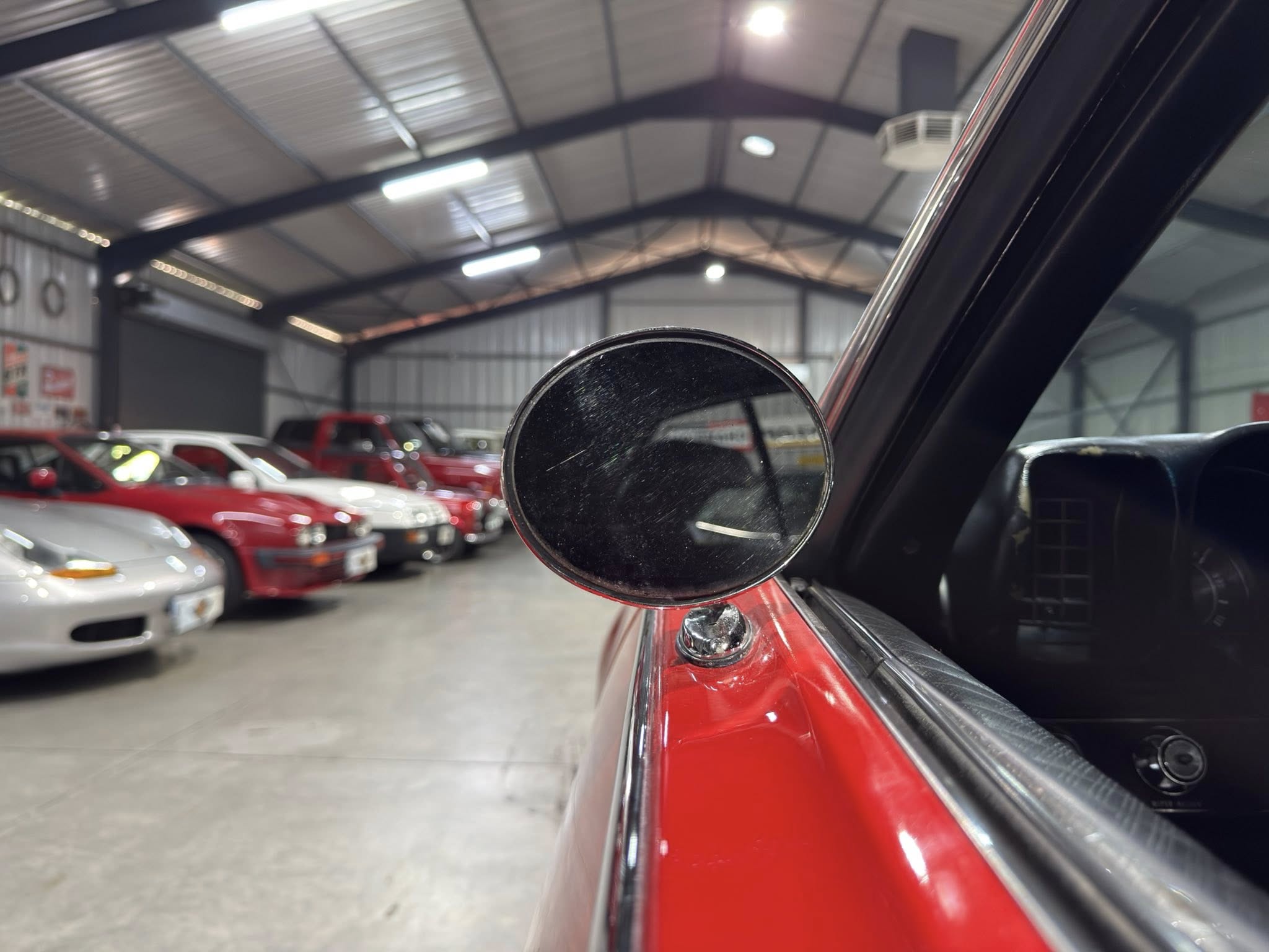 In a large indoor car showroom, a red car is visible with a round side mirror in the foreground.
