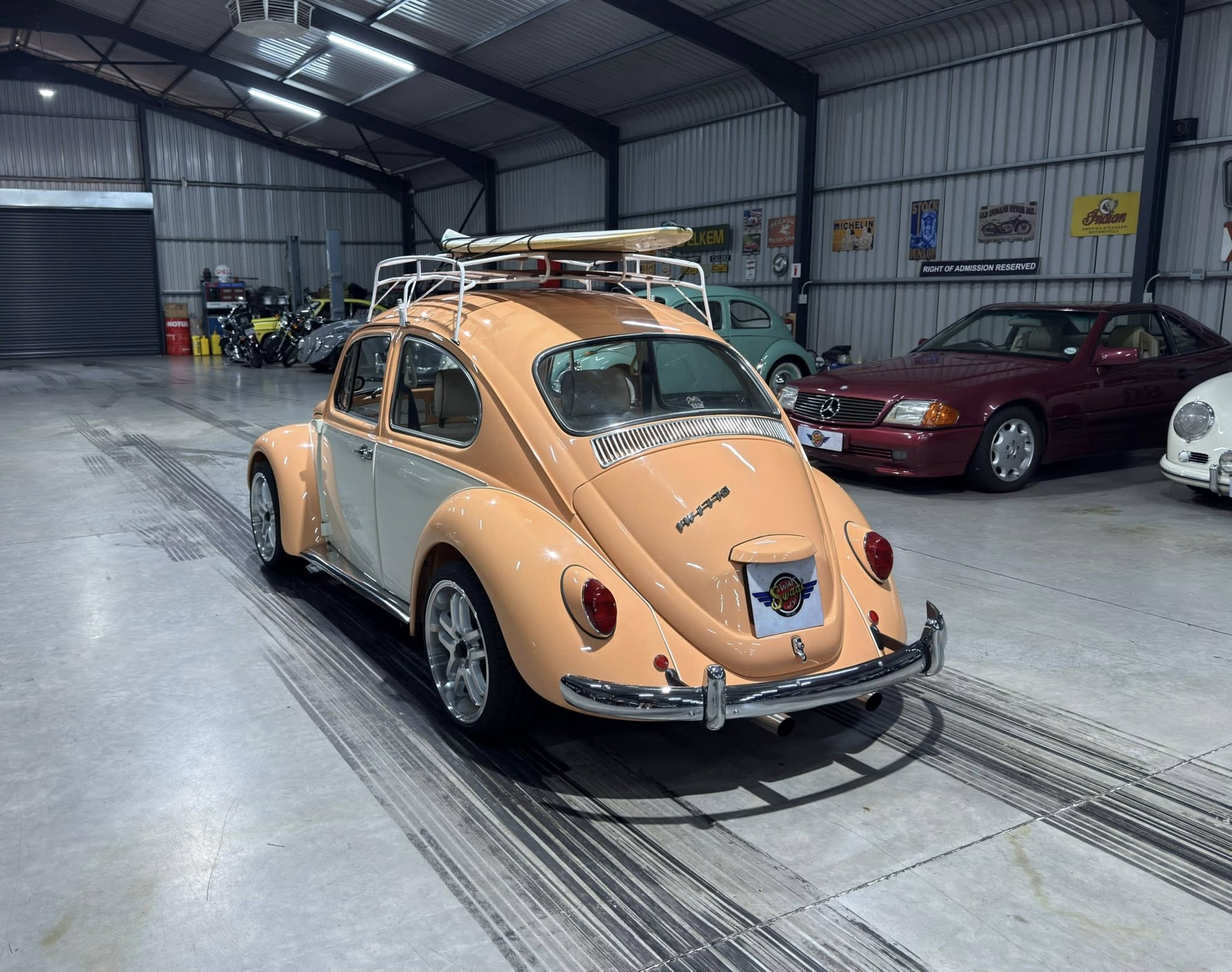Peach-colored vintage VW Beetle with a surfboard on its roof parked in a showroom beside other classic cars and bikes under a metal roof.