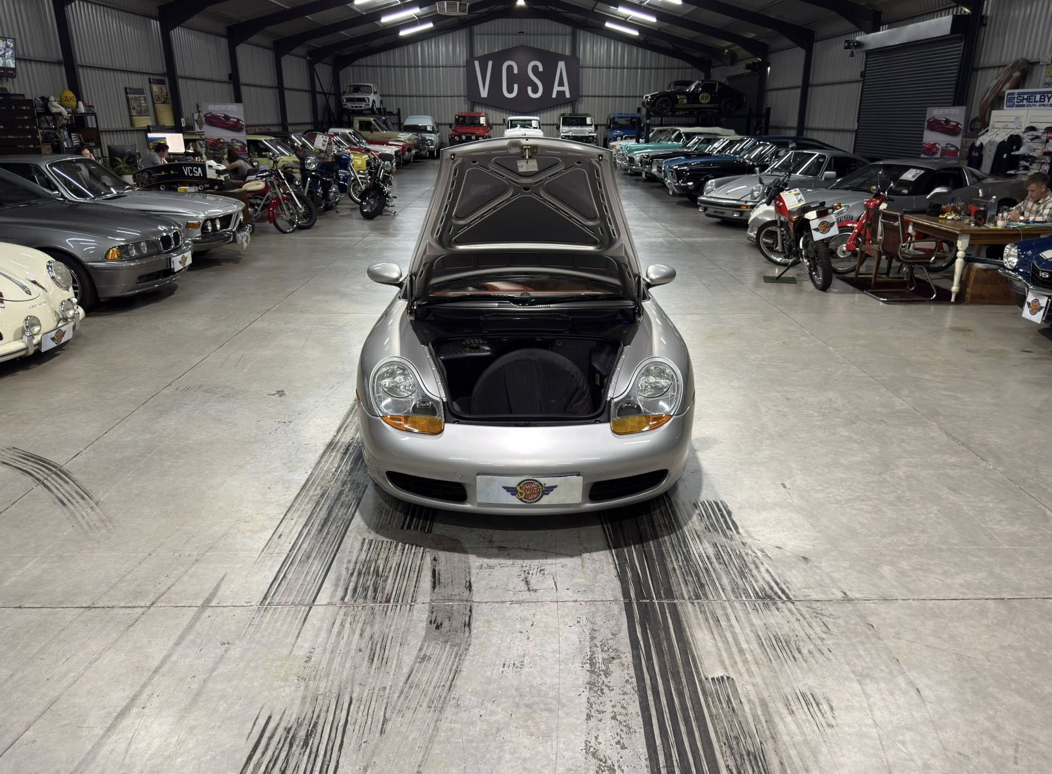 Open hood view of a silver sports car in a spacious classic car showroom, with rows of vintage cars and motorcycles on both sides and a 'VCSA' sign in the back.
