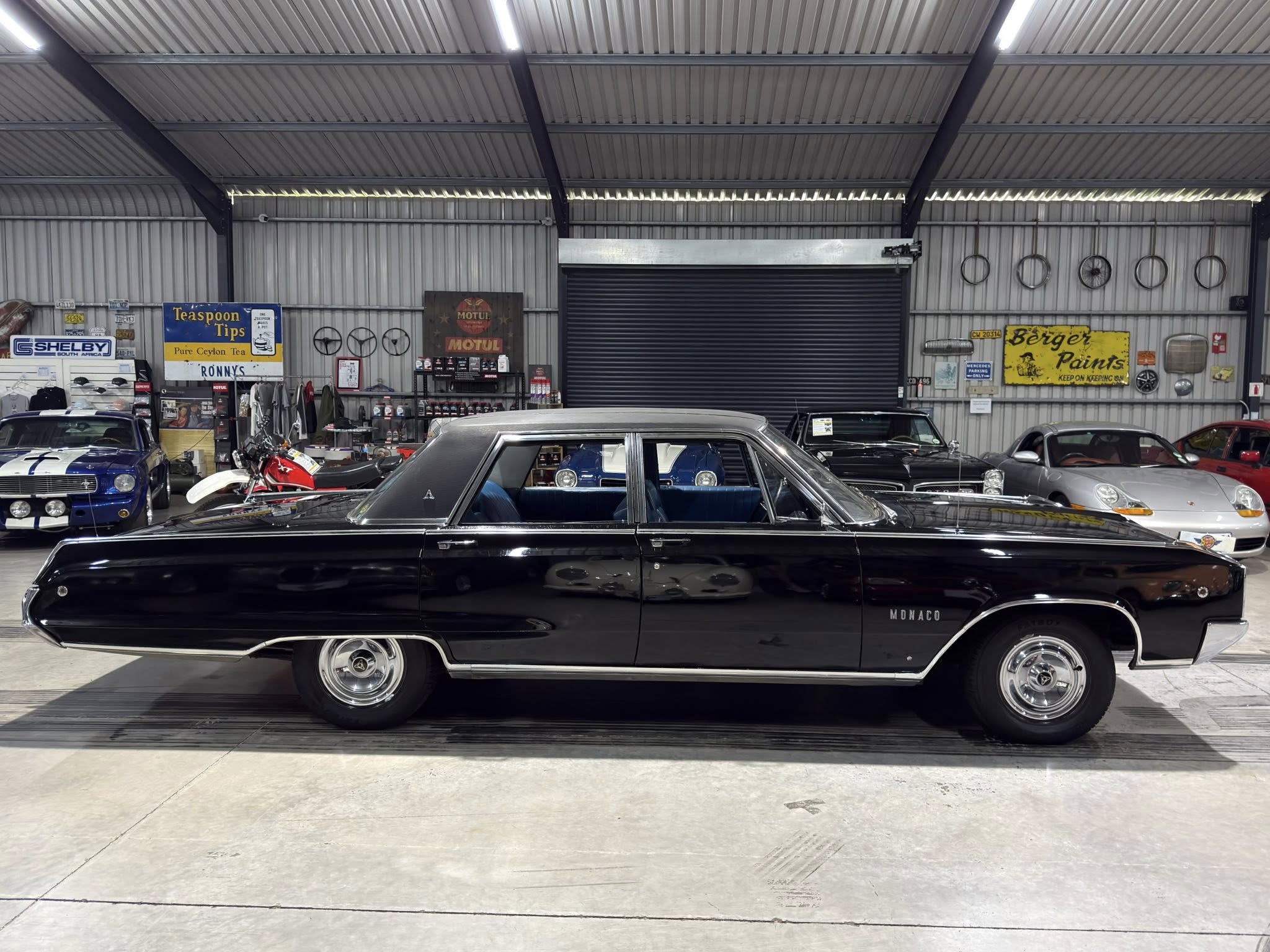 Inside a metal‑walled showroom with several classic cars, including a prominent black Mercedes in the foreground and a rolling door in the background