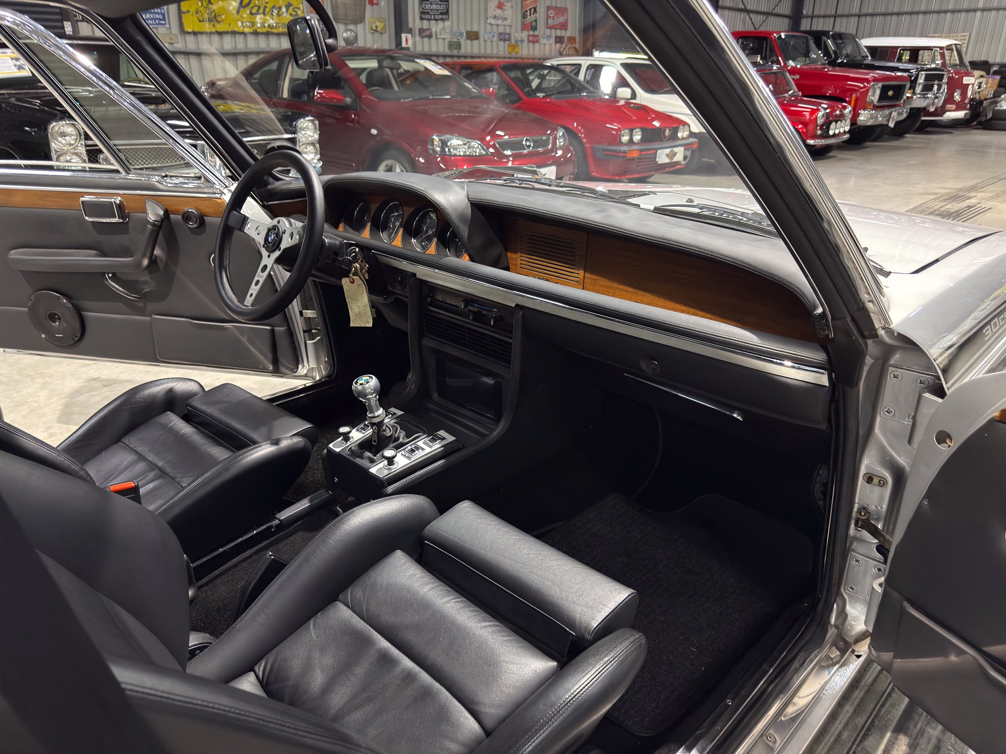Inside a vintage car with black leather seats, wood dashboard trim, and a gear shifter, shown in a showroom full of classic cars.