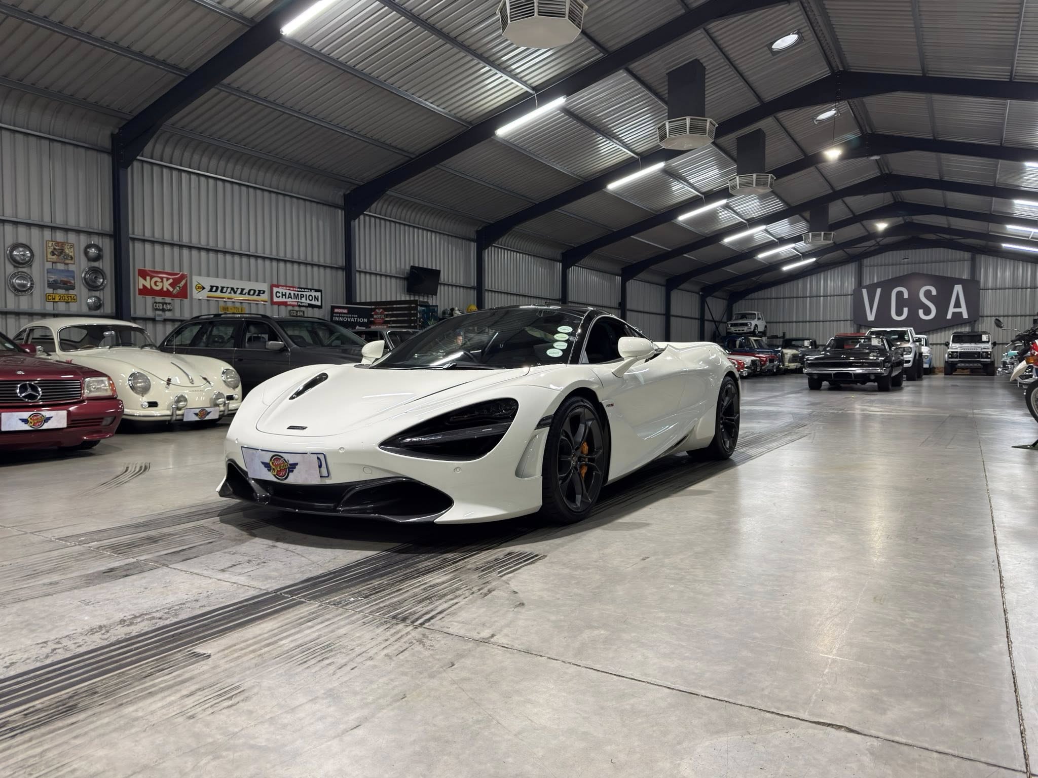 Indoor car showroom with a white sports car in the foreground and classic cars along the walls.