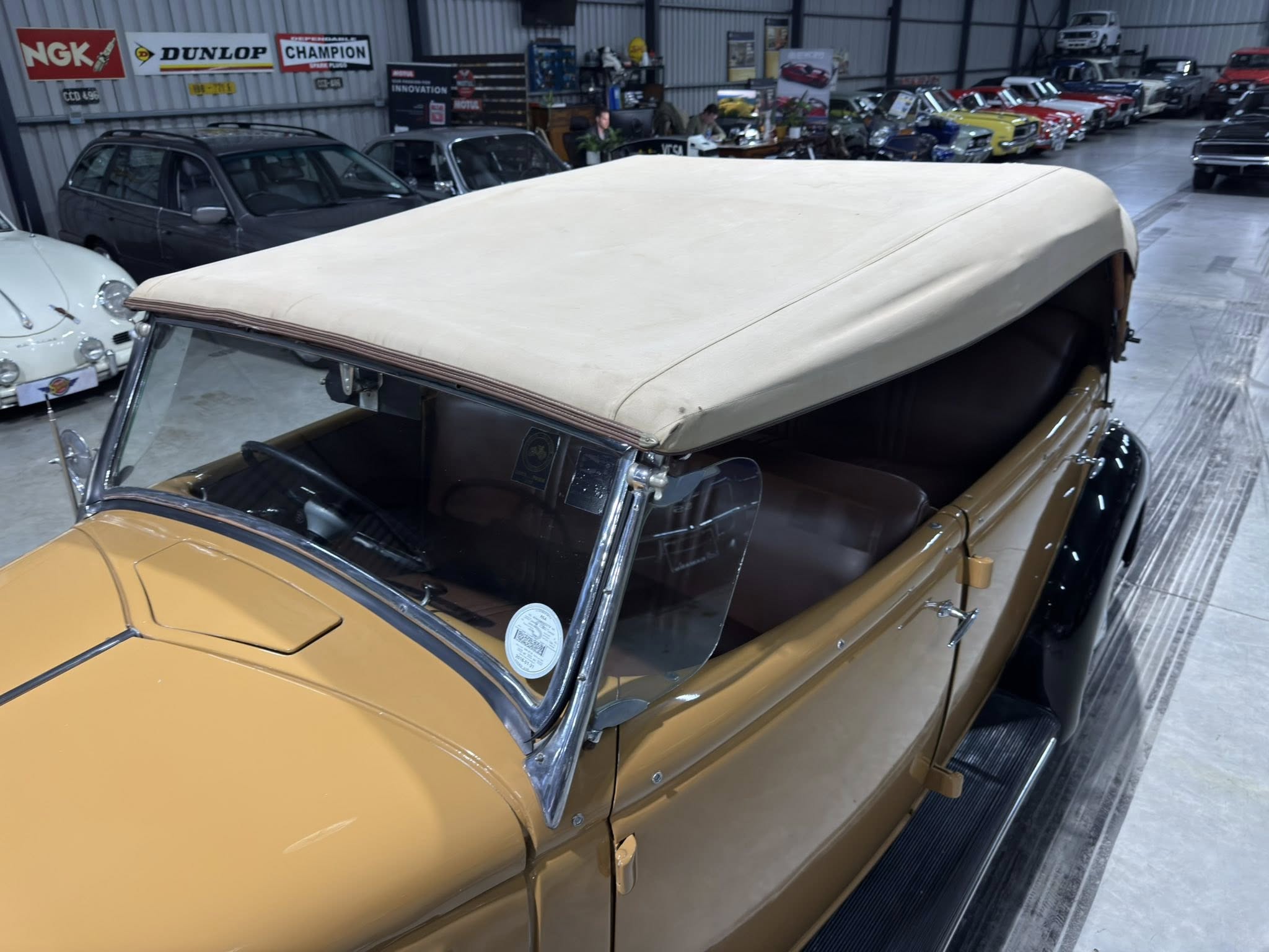 Vintage tan convertible with a raised white top in a car showroom, surrounded by classic cars and automotive banners in the background.