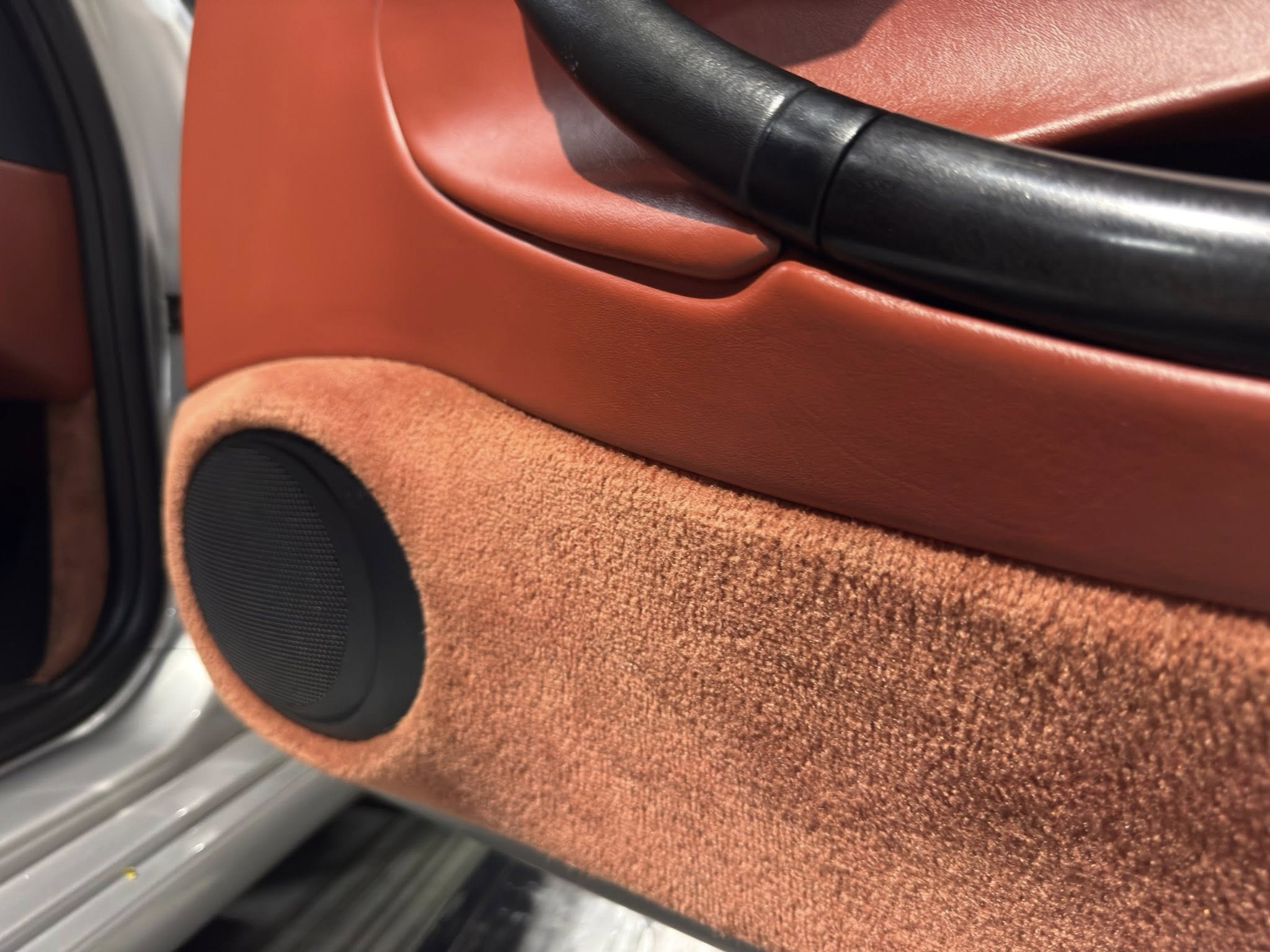 Close-up of a car door interior in red leather with a black armrest and a round speaker in the lower door panel.