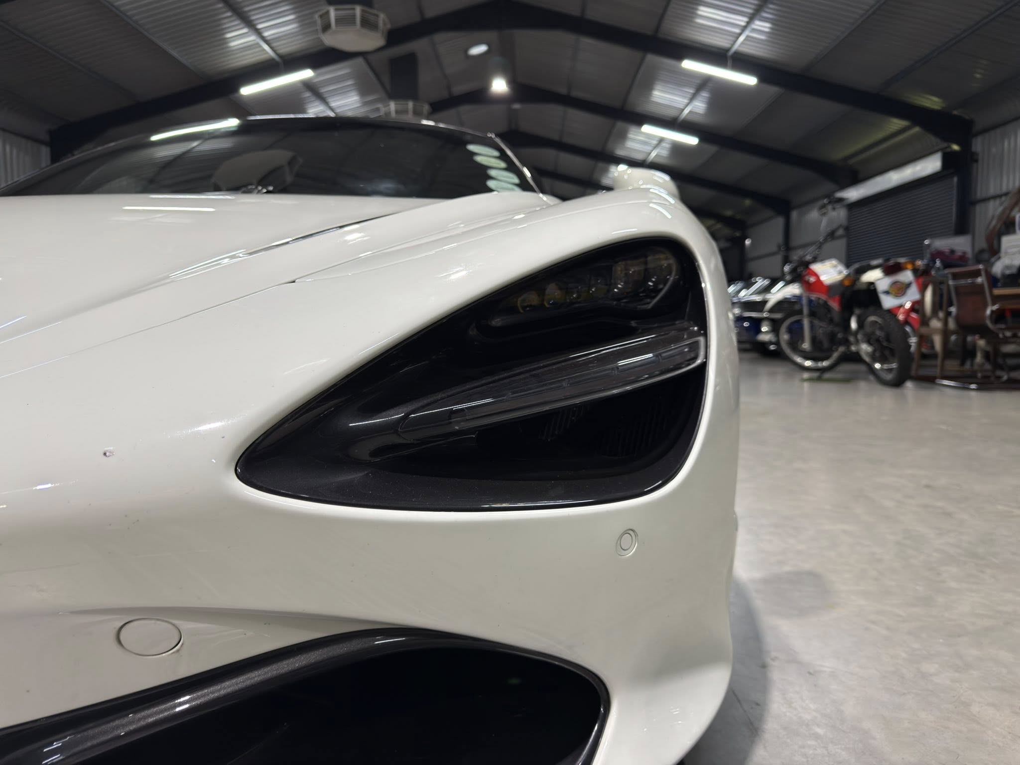 Close-up of the front left side of a white car in a showroom, with motorcycles blurred in the background.