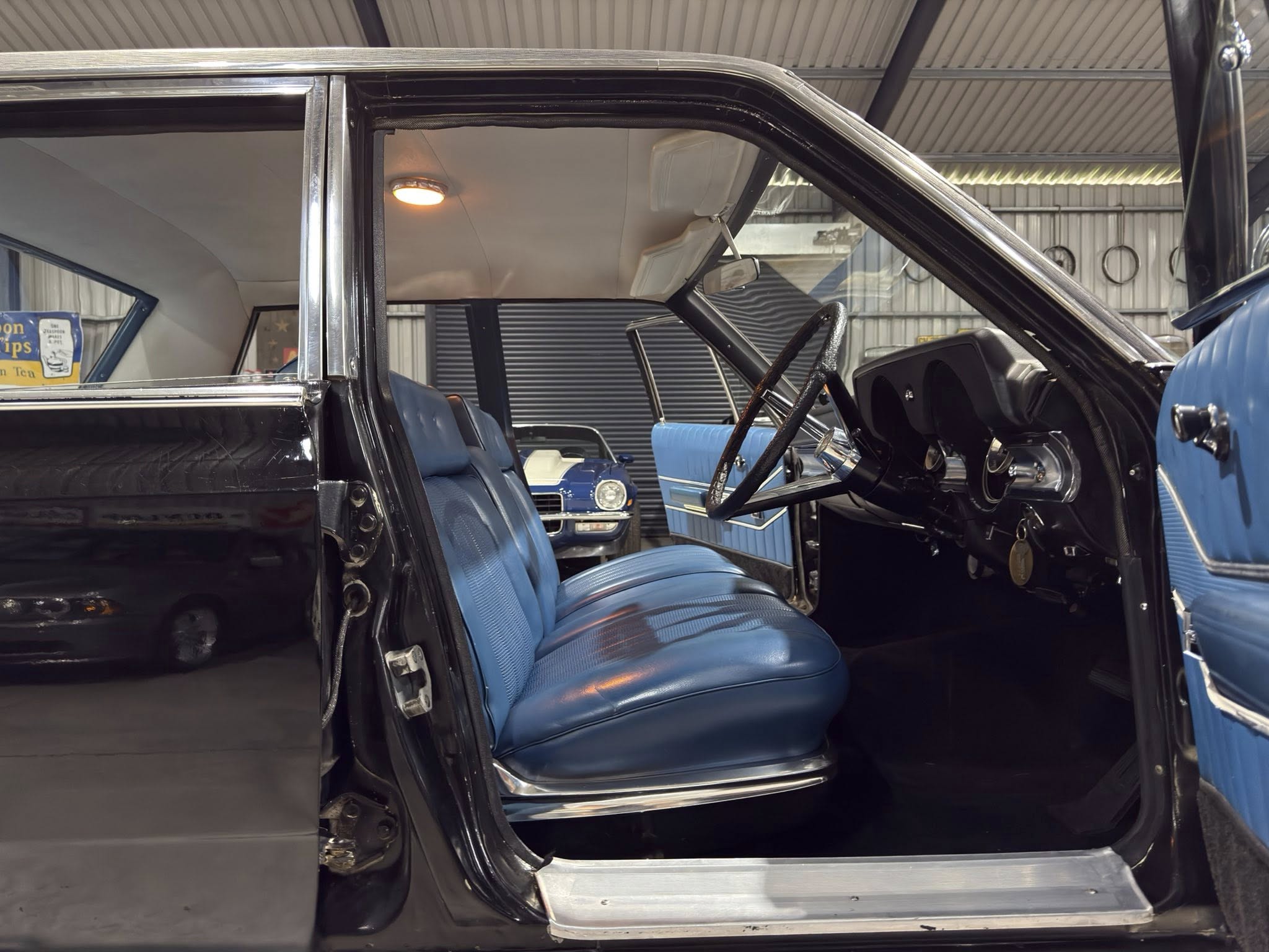 Interior of a vintage black car with blue vinyl seats and the driver's door open, showing the steering wheel and dashboard.Interior view emphasizes seating and cabin layout of classic car.