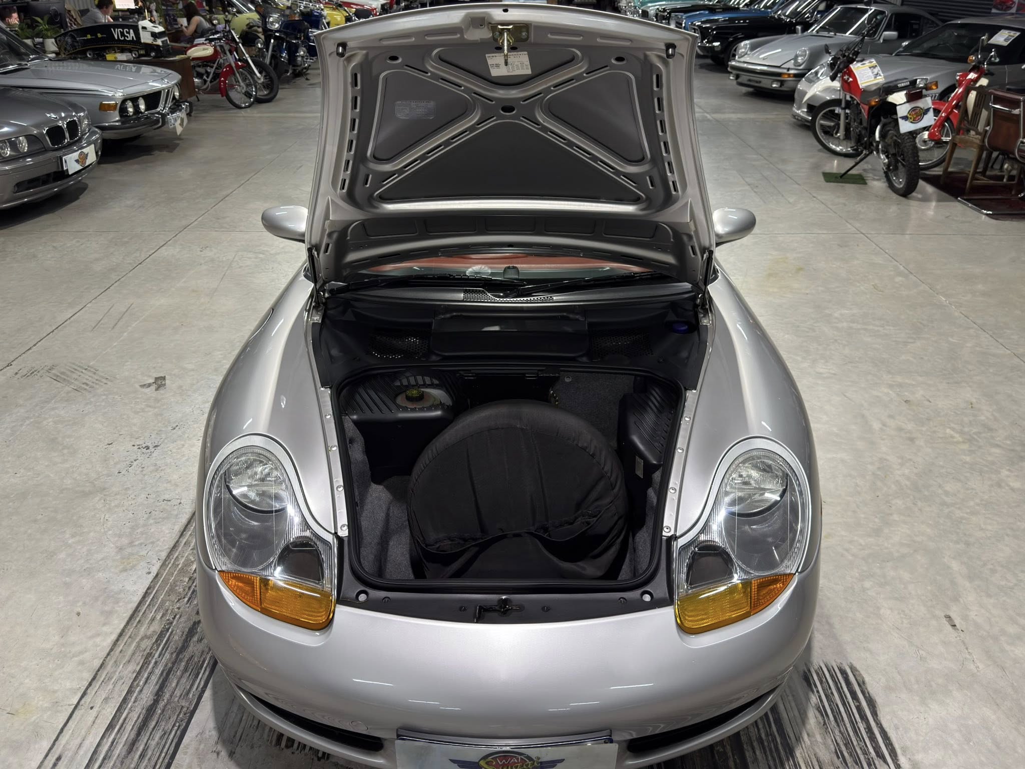 Front view of a silver sports car with its hood open inside a showroom, revealing the engine bay and tires.