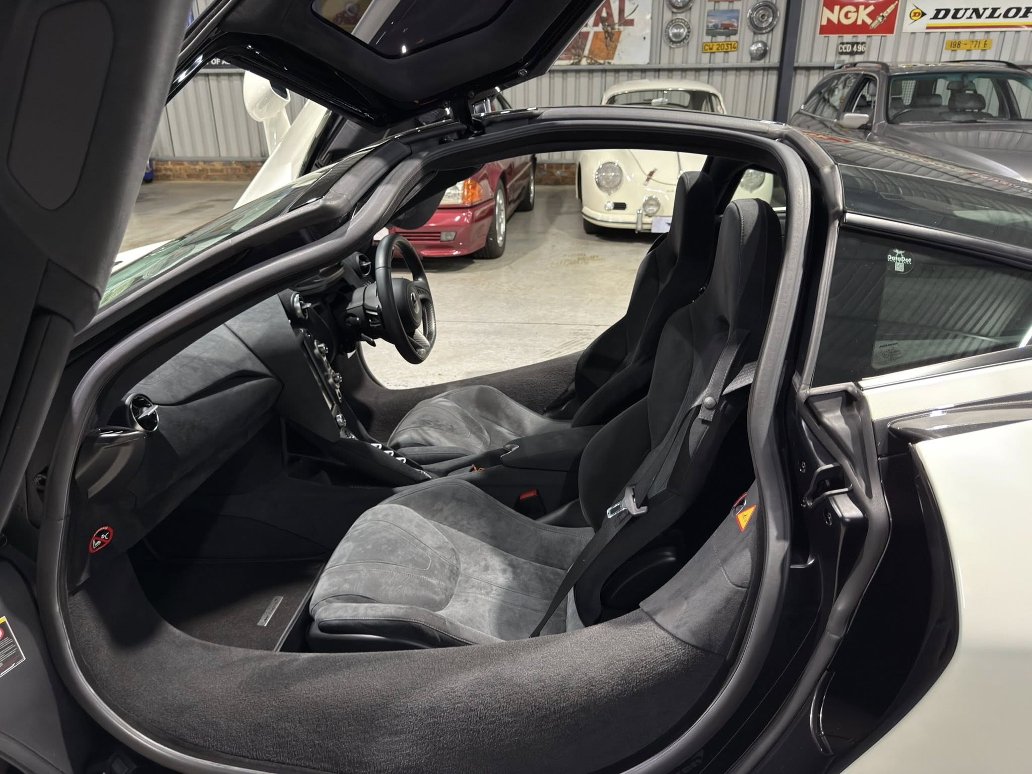 Interior of a sleek sports car with gull-wing door raised, black seats, and dashboard visible; showroom with vintage cars in the background.