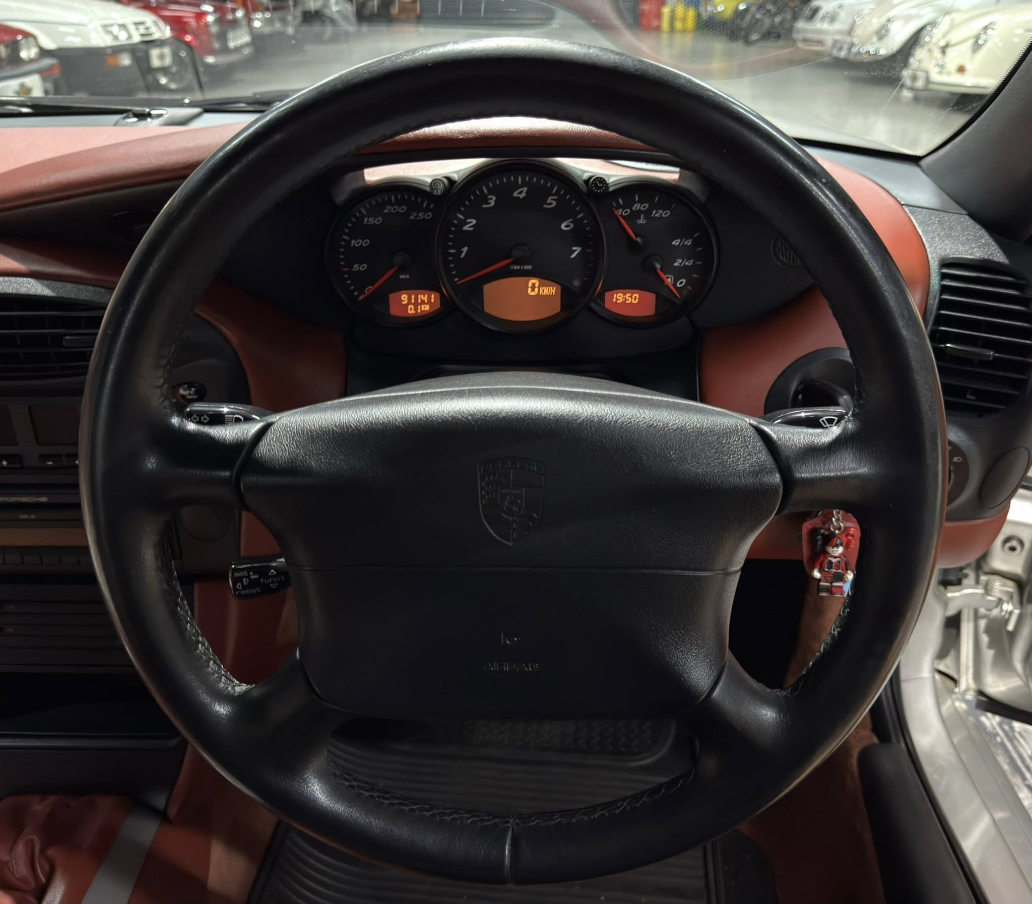 Interior view of a car dashboard from the driver's seat, with a black steering wheel and orange-lit instrument cluster showing speed and rpm gauges in a red-brown interior.