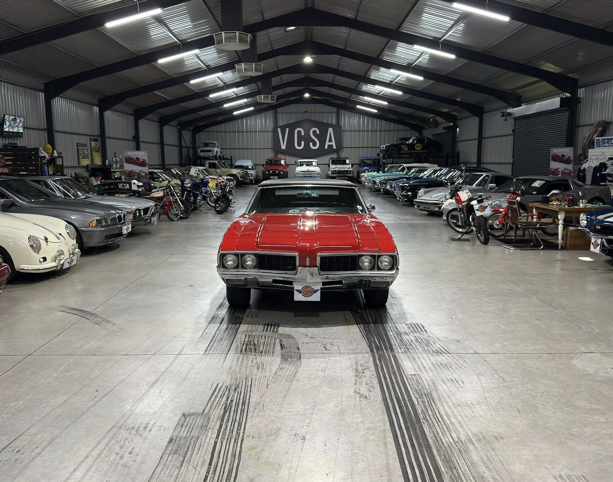 Indoor car showroom filled with vintage cars, red car centered in foreground, rows of classics along both sides, VCSA sign on back wall.