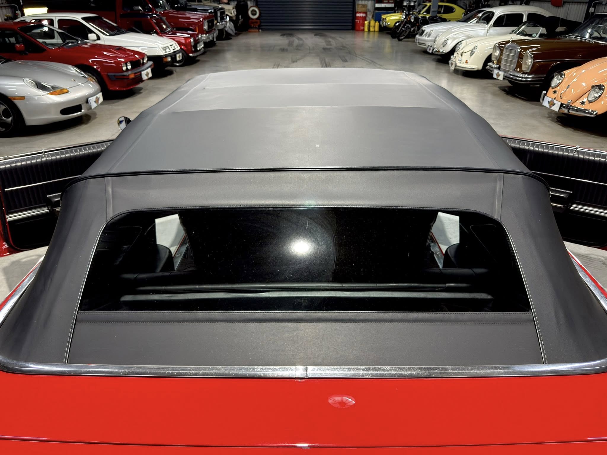 Back view from a foreground convertible looking over its soft top at a showroom filled with vintage cars along both walls.