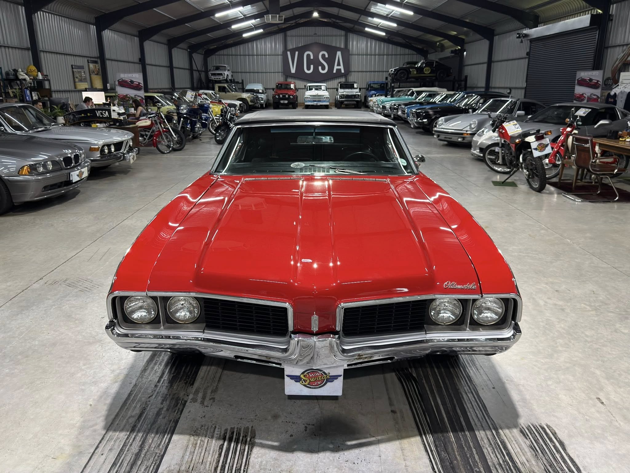 Red classic car in the foreground inside a large vintage car showroom with rows of classic vehicles and a VCSA sign in the back gallery window.