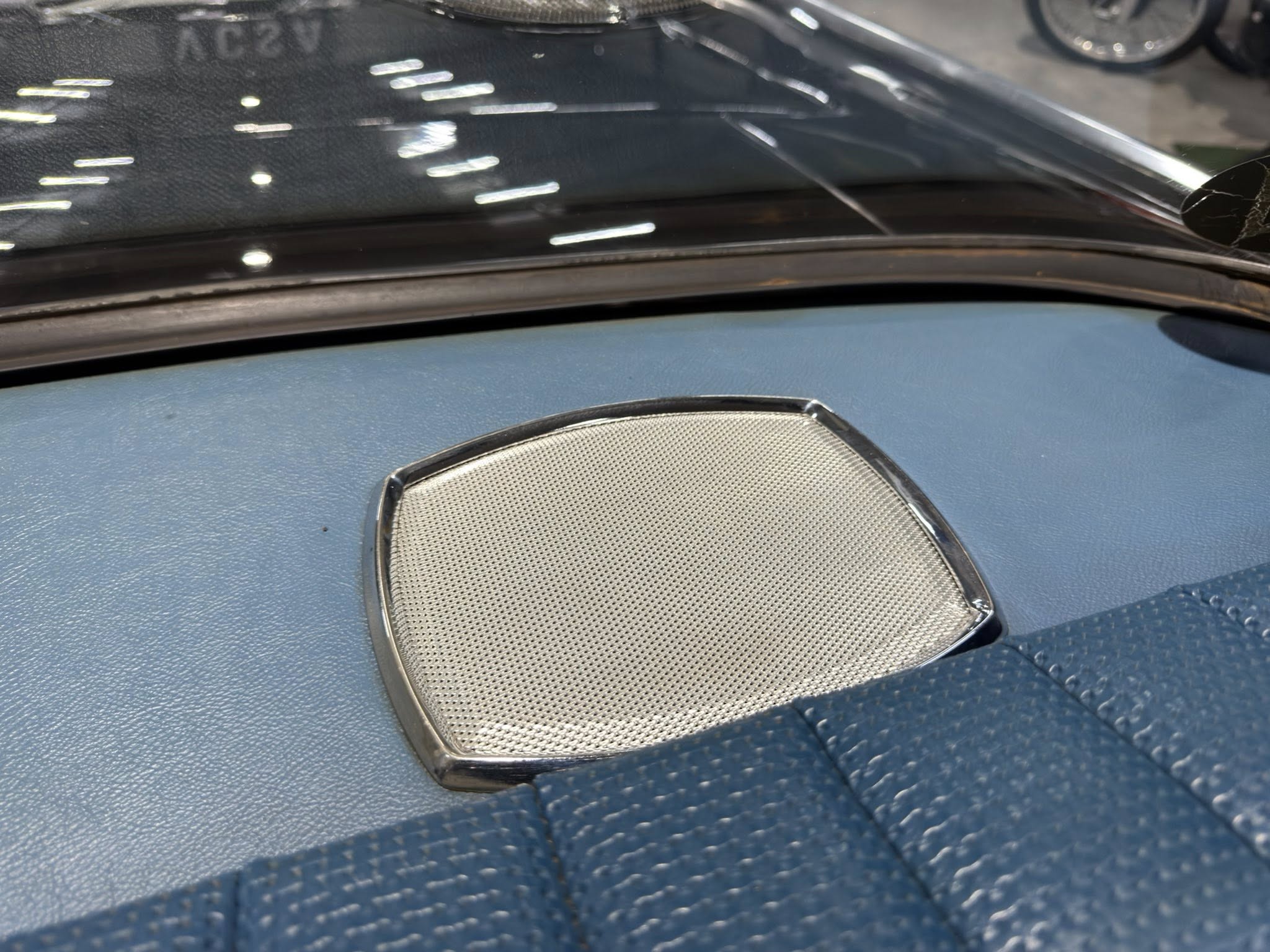 Close-up of a blue car dashboard with a beige perforated speaker grille in the foreground and a cracked windshield edge on the right.