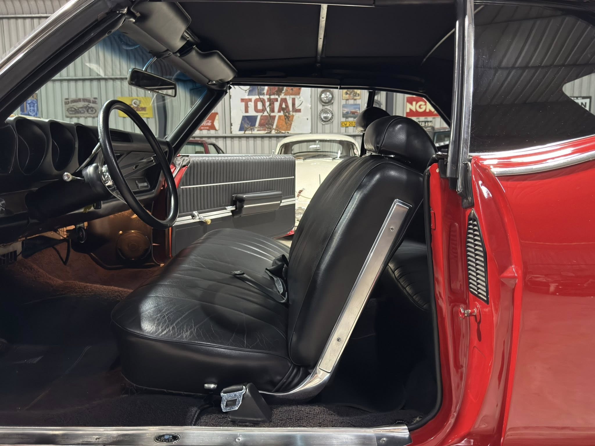 Inside view of a vintage red car showing black leather seats, steering wheel, and open door in a garage showroom.