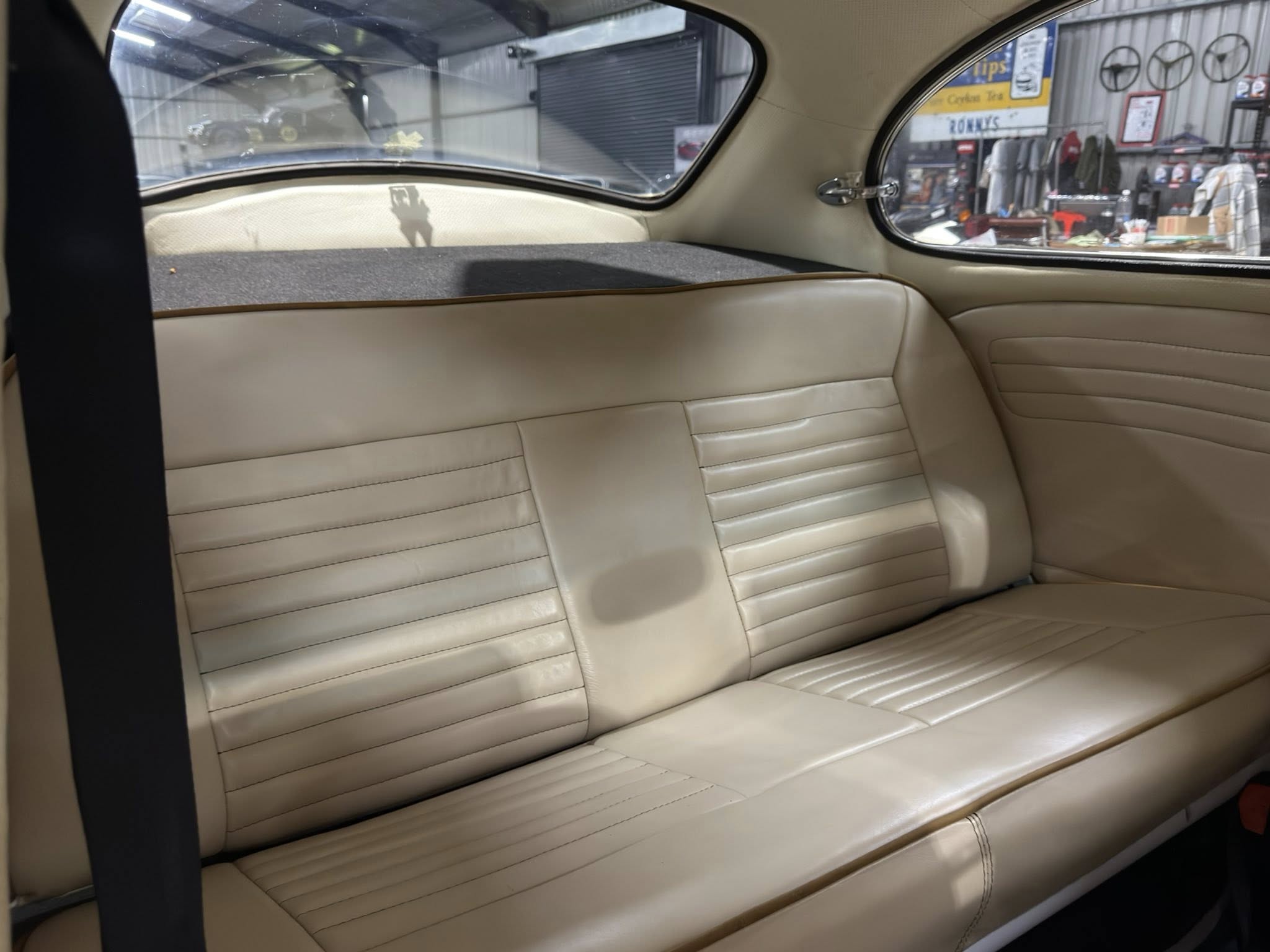 Back seat interior of a beige classic car, showing horizontal stitched upholstery and rear window view of a cluttered workshop.