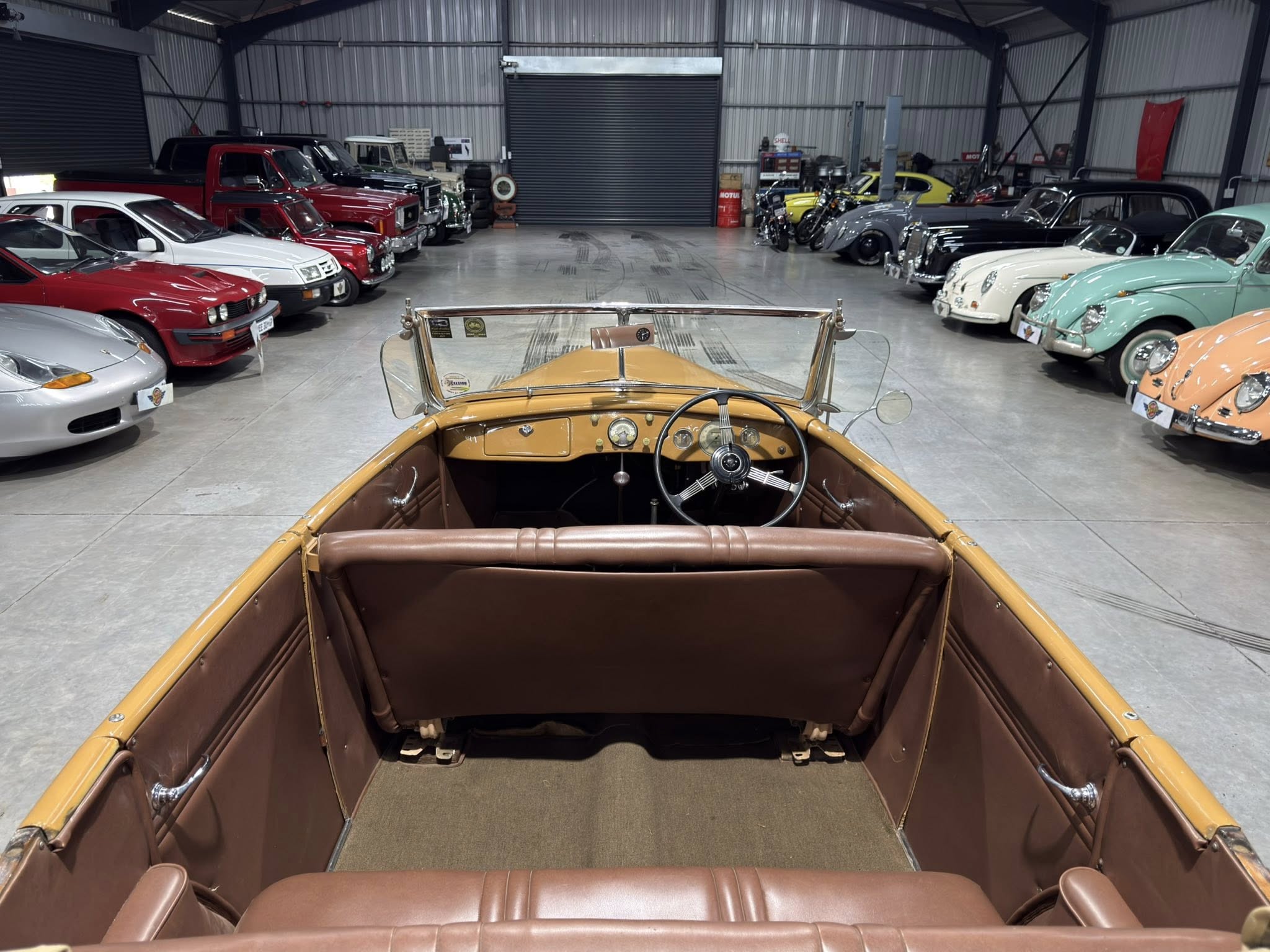 View from the driver's seat of a tan vintage convertible, looking down the car’s interior toward a showroom filled with classic cars on both sides.