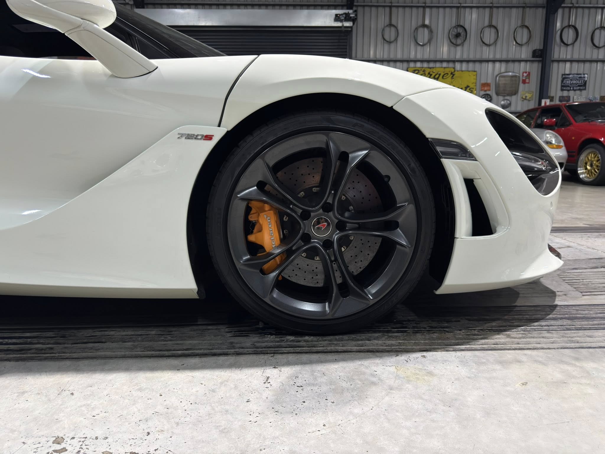 Close-up of a white sports car's front left wheel with orange brake calipers visible through a grey alloy rim, Ferrari 488 S badge on the fender, in a workshop.