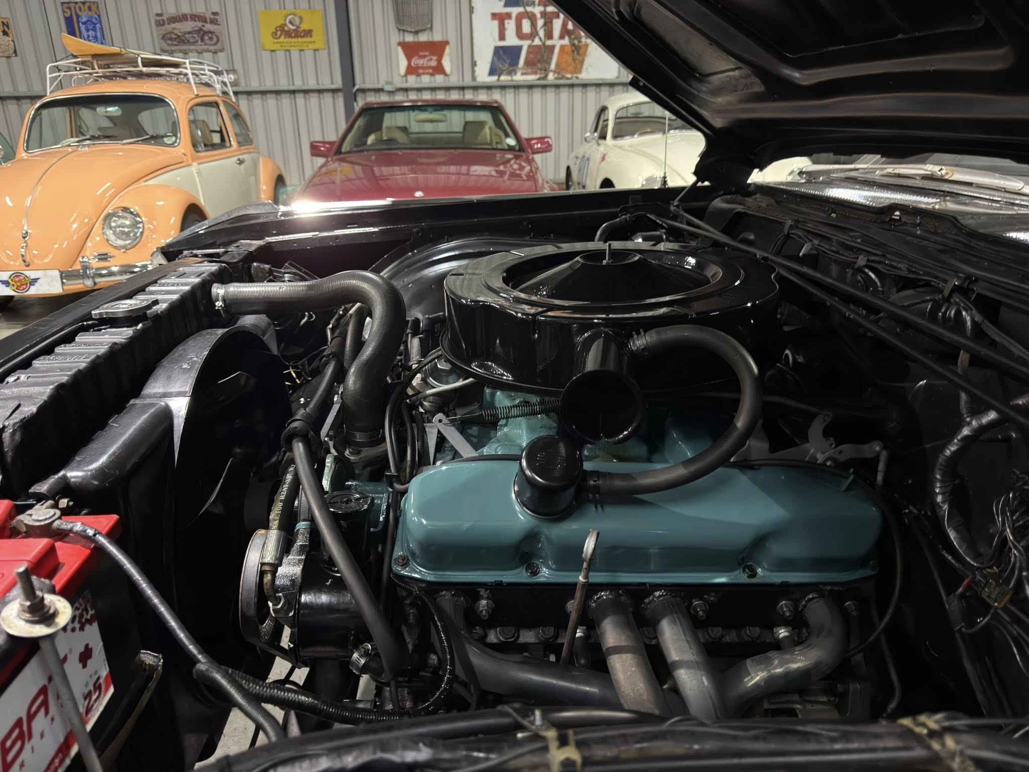 View from inside a car’s open hood showing an engine bay, with vintage orange and red classic cars on display in a garage in the background.