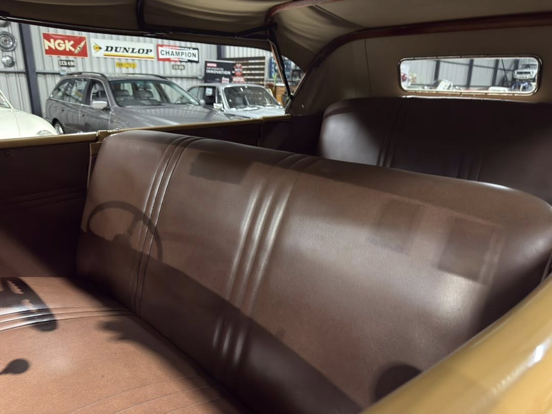 Rear interior of a vintage car with brown leather seats and a window view of a garage yard with classic cars and signboards outside of the shop.