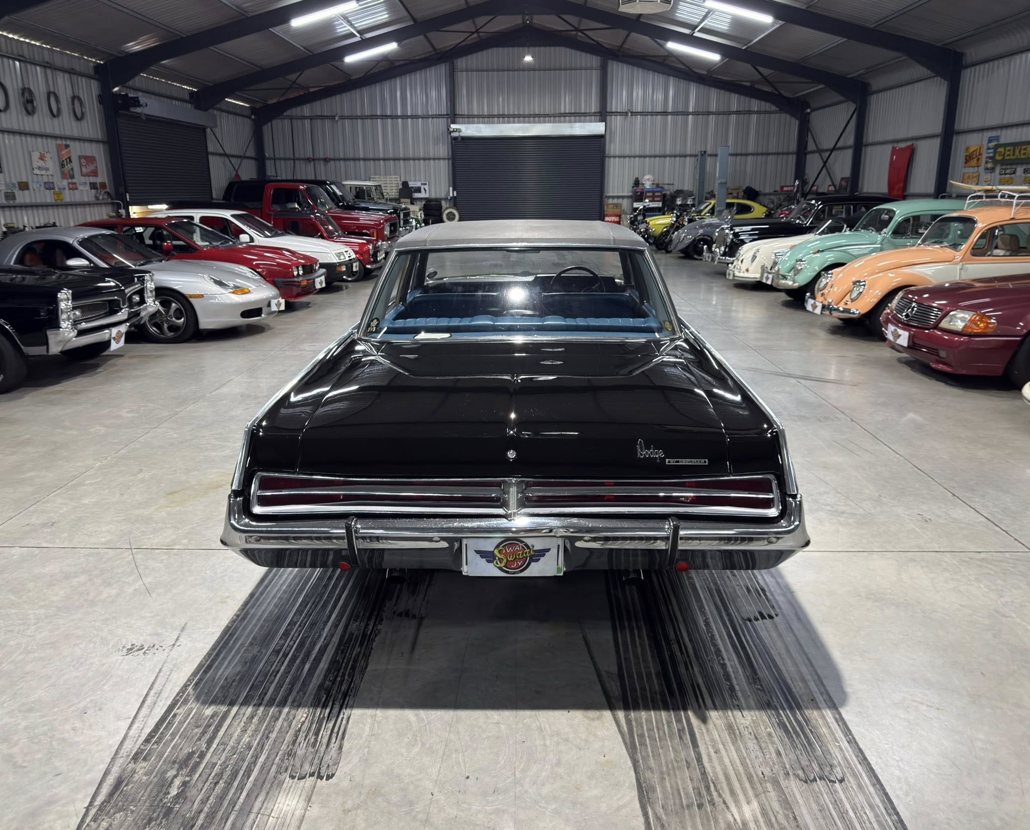 Indoor showroom full of classic cars, viewed from behind a black vintage car at the center aisle.
