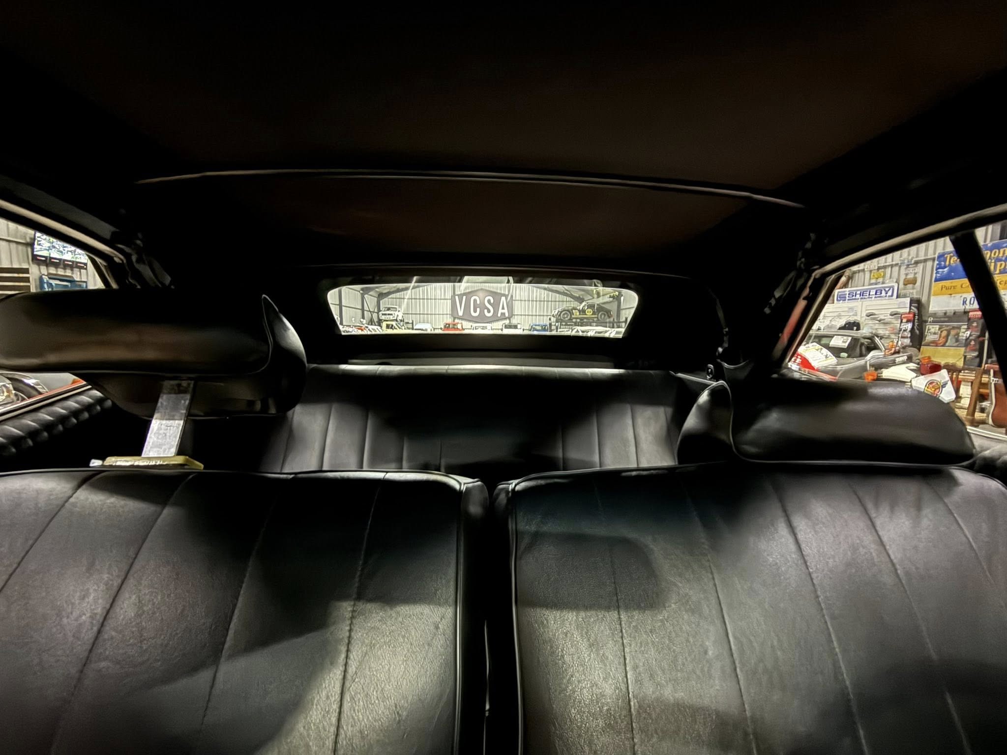 Interior of a car with black leather seats, photographed from the back seat; through the rear window you can see a garage yard and a 'VCSA' sign with parked cars outside