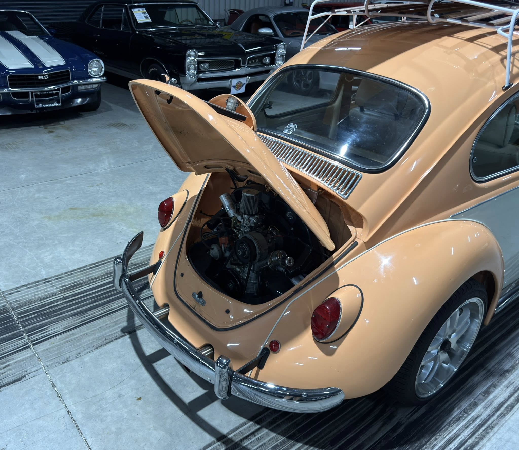 Peach-colored classic car with its rear hood open, engine area visible, in a showroom with other vintage cars in the background.