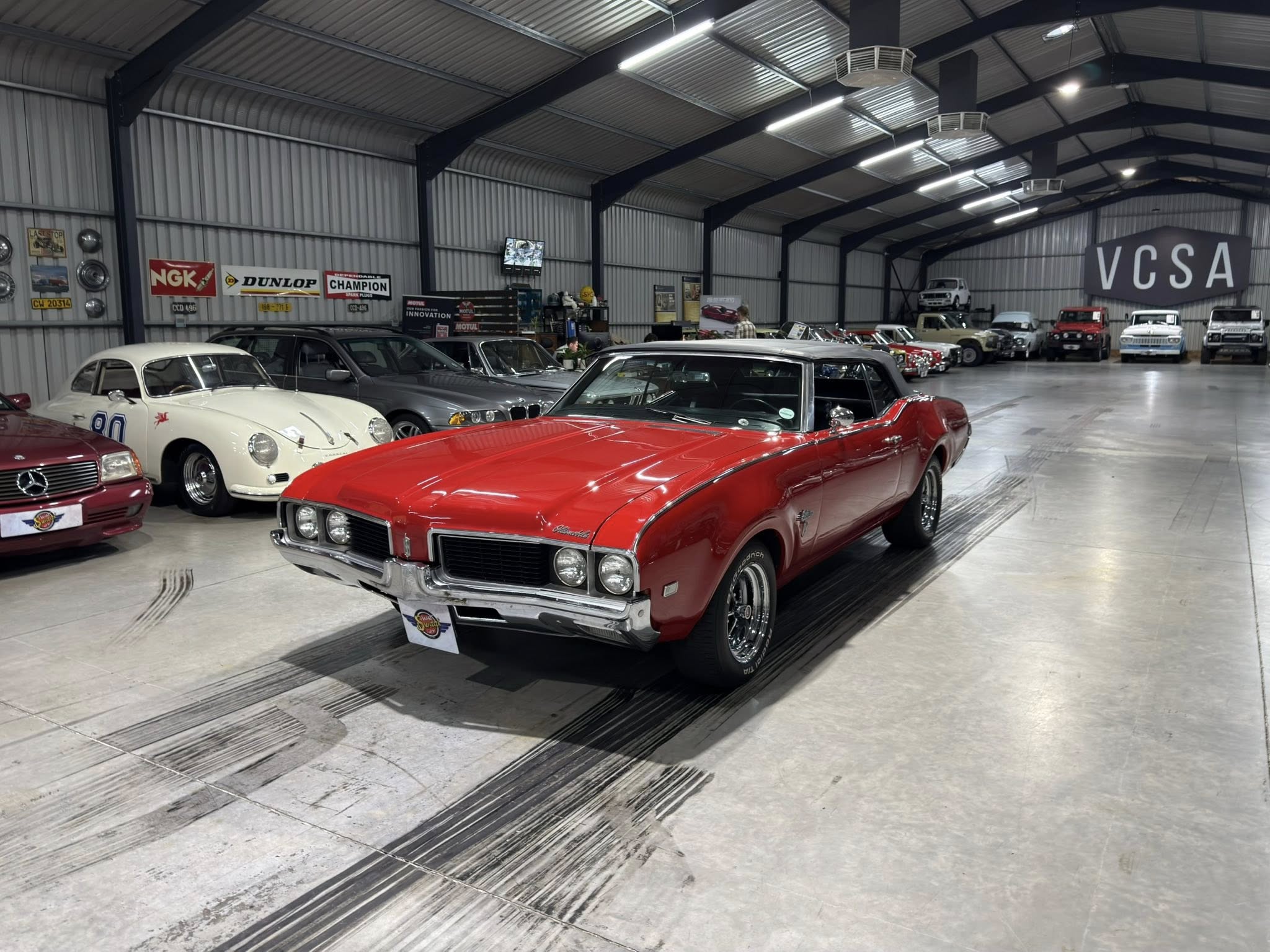 Indoor showroom full of classic cars, with a bright red convertible in the foreground and other vintage vehicles along the sides.