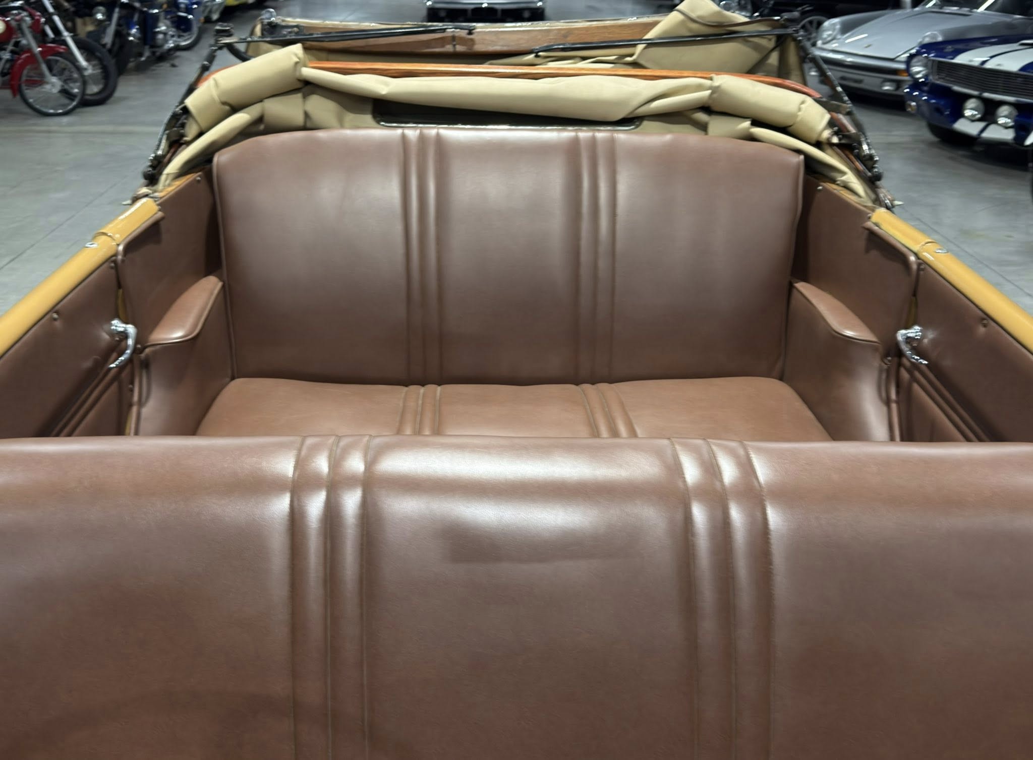 Rear seating area of a vintage brown leather convertible with the top folded back; showroom with other classic cars in the background.