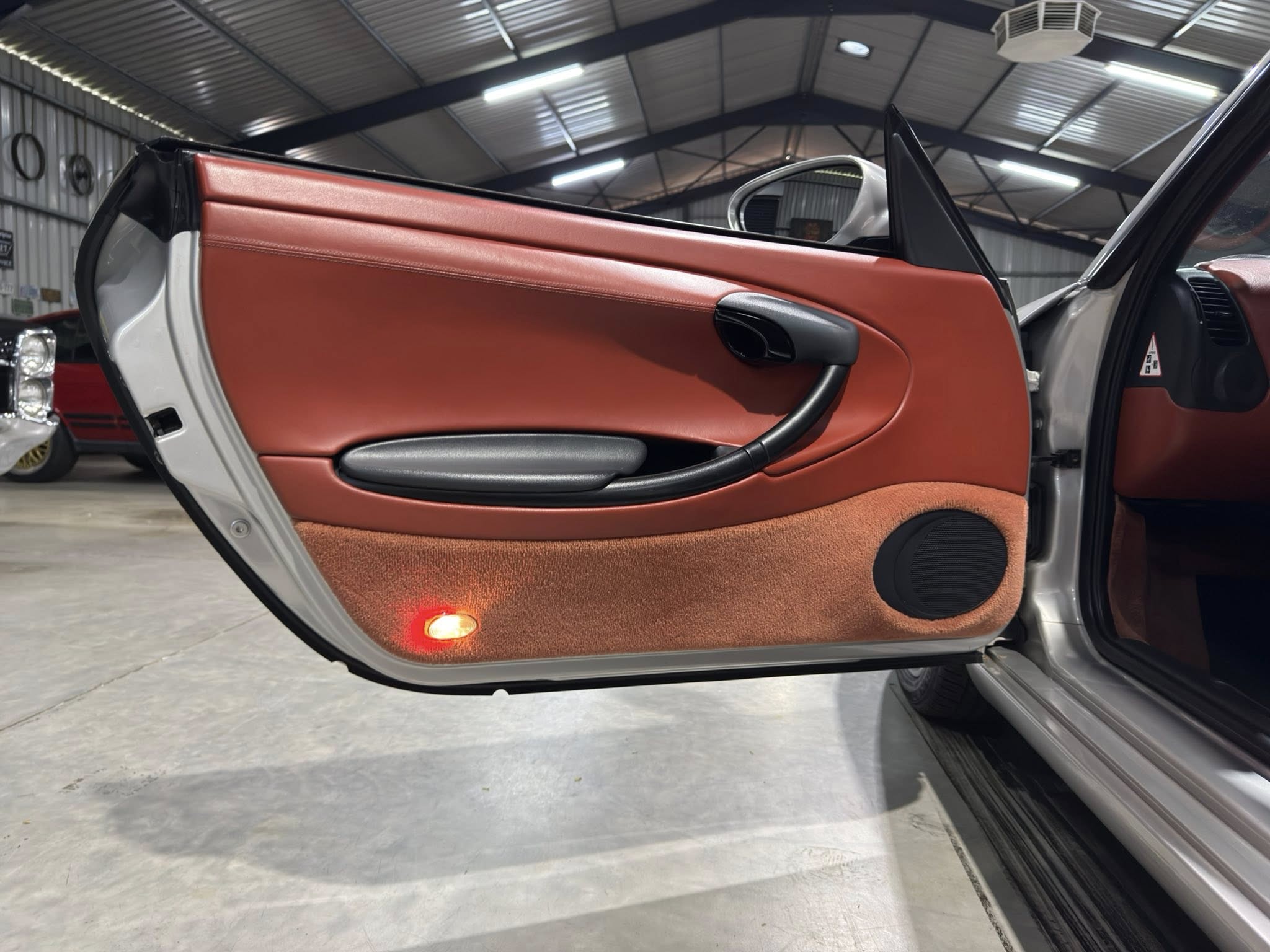 Open car door showing red leather interior panel, black armrest, and a red courtesy light near the bottom in a garage.
