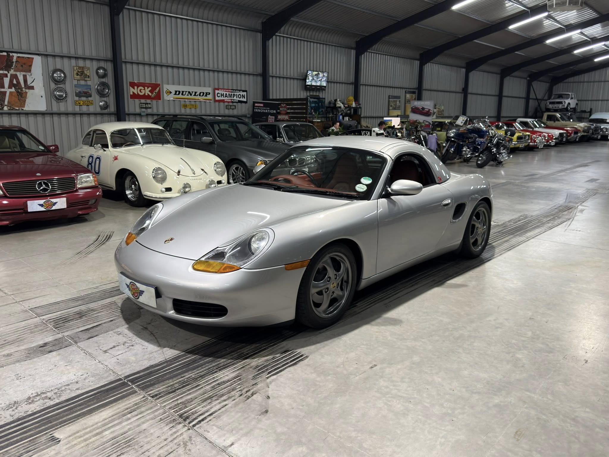 Indoor showroom full of classic cars, including a silver convertible in the foreground and a white vintage coupe nearby.