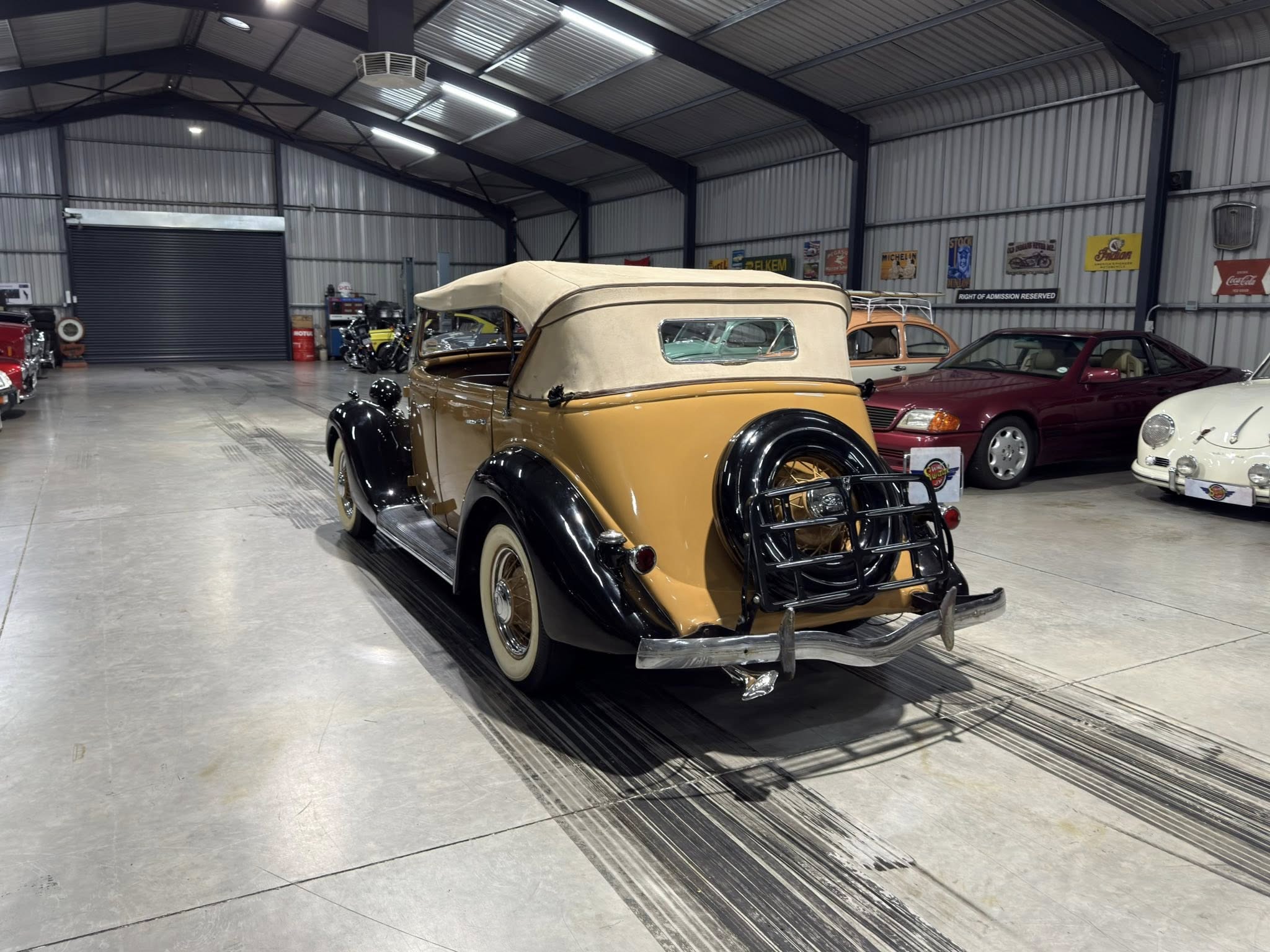 Indoor showroom of classic cars; a tan vintage convertible dominates the foreground with other antique cars along the back wall.
