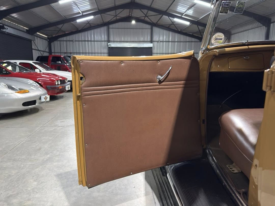 Open vintage car door showing brown door panel and interior seating in a garage setting with other cars in the background, indoor lighting.