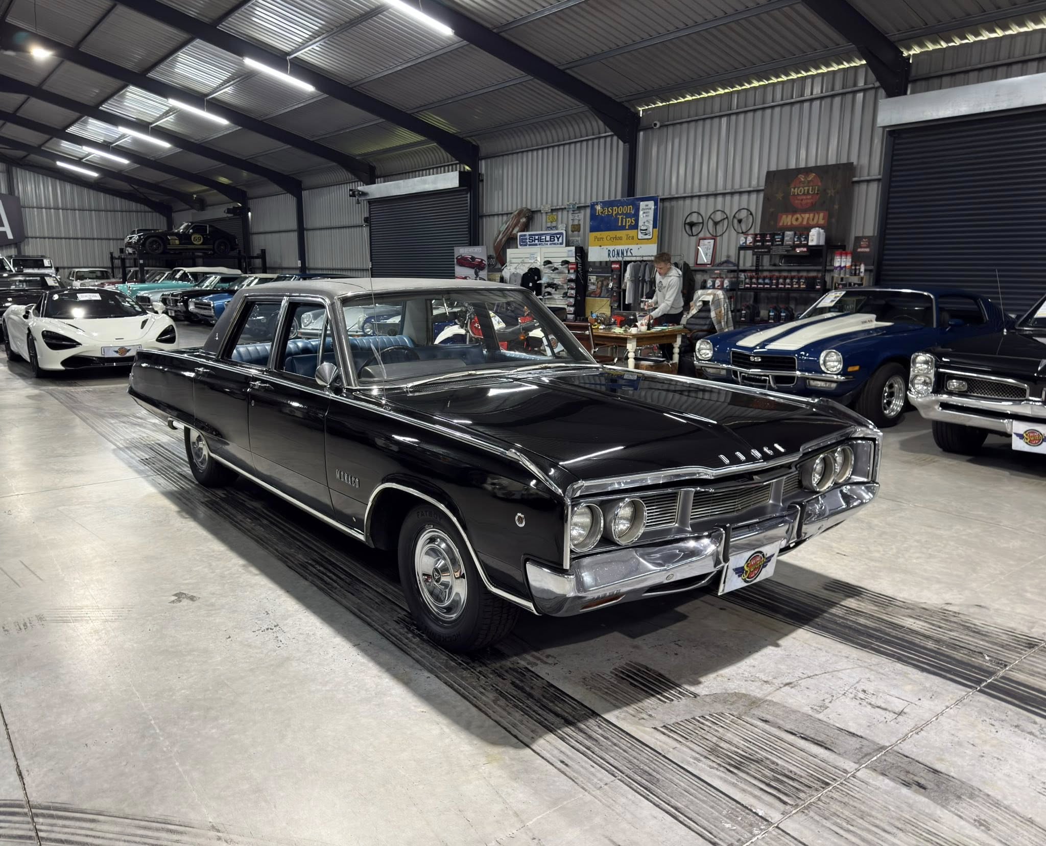 Indoor showroom of vintage cars with a prominent black classic car in the foreground and more classics lined up in the background.