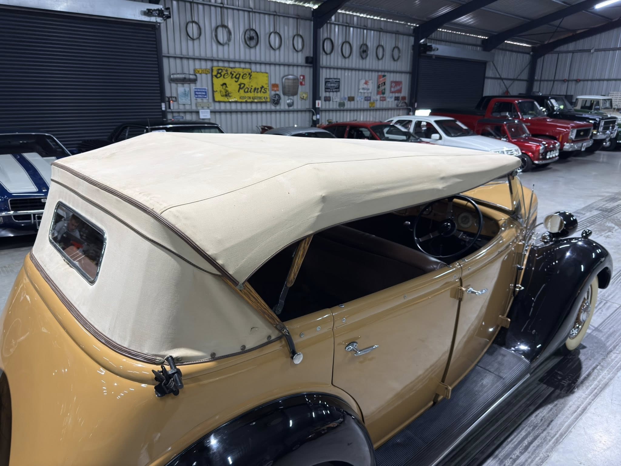 Vintage tan convertible in a garage showroom surrounded by other classic cars and retro signs on the wall