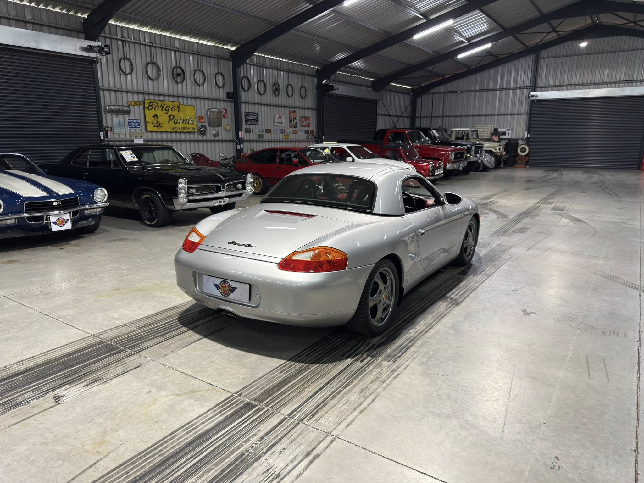 Interior of a large metal warehouse turned into a classic car showroom, with a row of vintage cars along the wall and a silver Porsche in the foreground.
